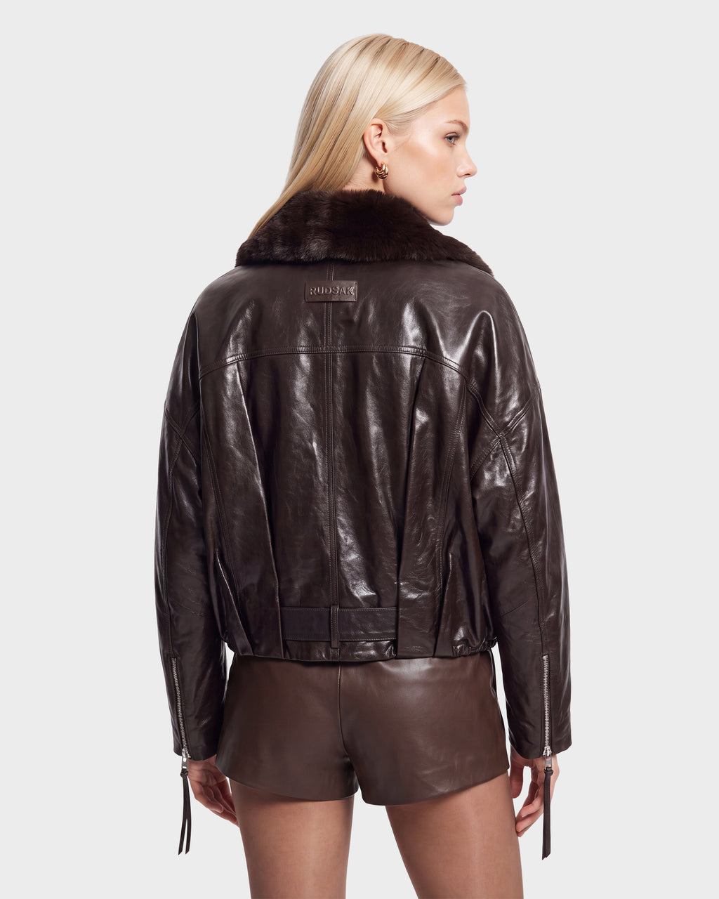 A woman with long blonde hair models the Rudsak AVITA - 8126938 Espresso, a dark brown leather jacket with a fur collar and matching shorts, standing against a plain light background, facing away and glancing to the side.