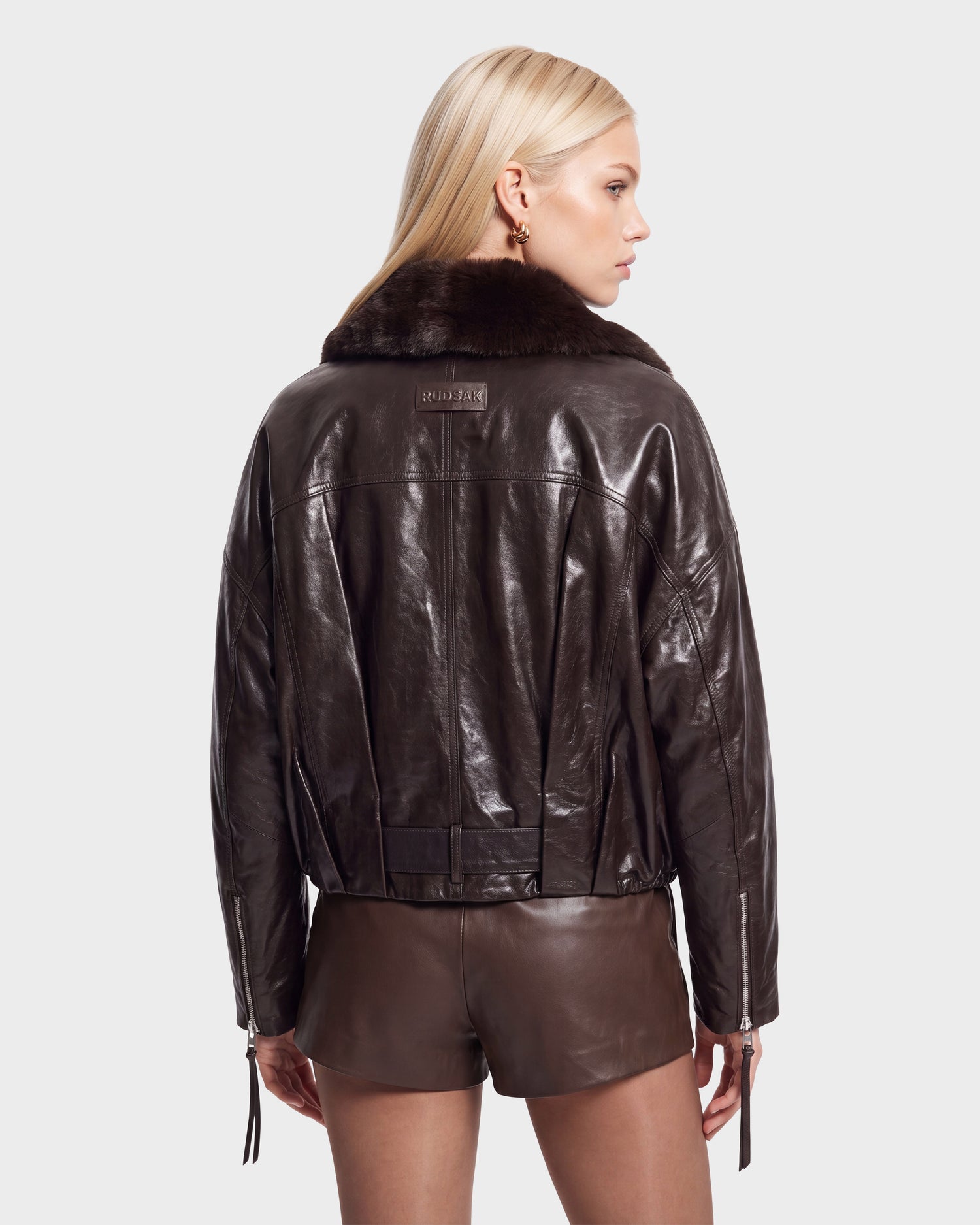 A woman with long blonde hair models the Rudsak AVITA - 8126938 Espresso, a dark brown leather jacket with a fur collar and matching shorts, standing against a plain light background, facing away and glancing to the side.