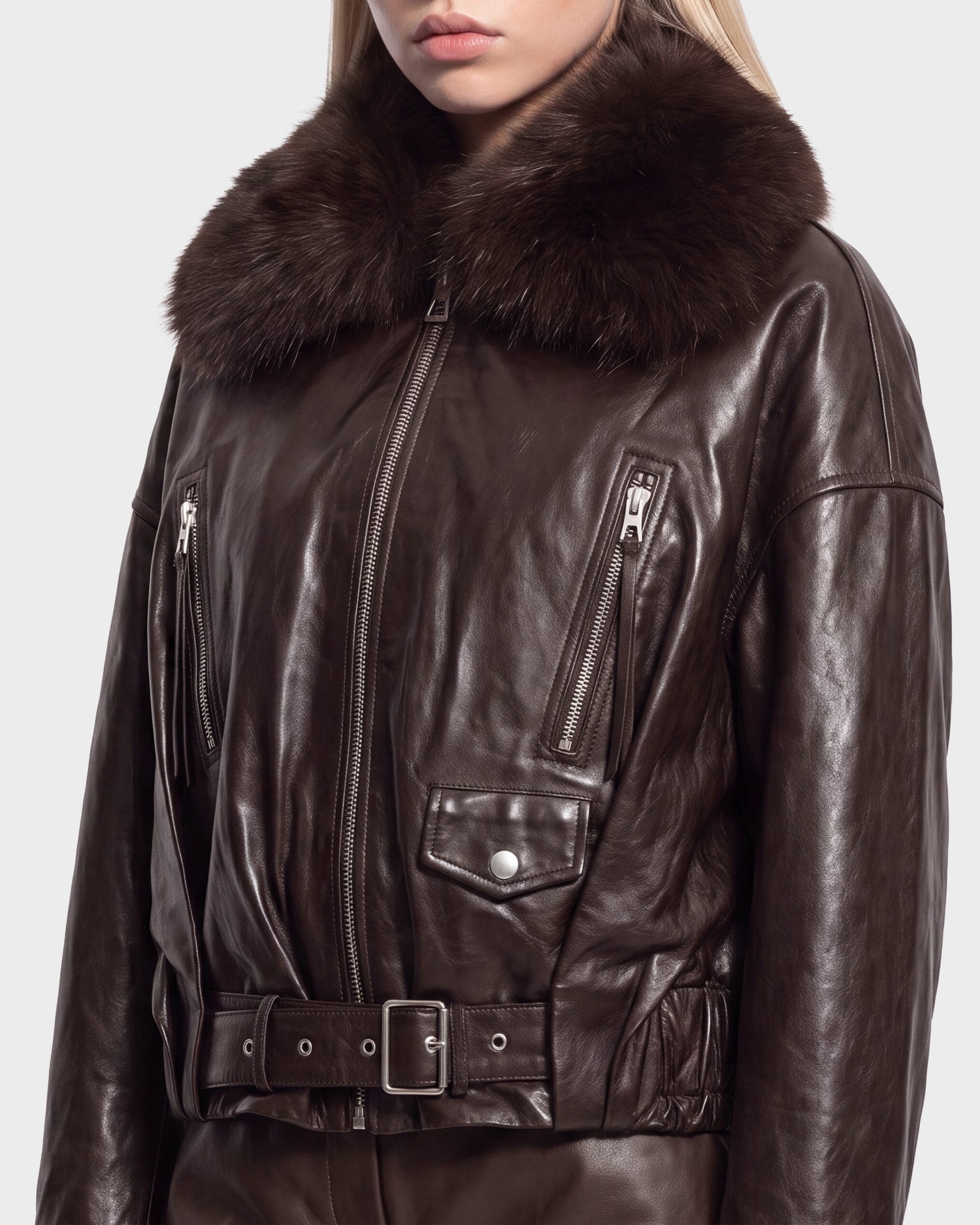 A woman models the Rudsak AVITA - 8126938 Espresso jacket, a dark brown leather piece featuring a fur collar, zippered chest pockets, a belted waist, and metallic snap accents. The image is cropped above her mouth and at the hips.