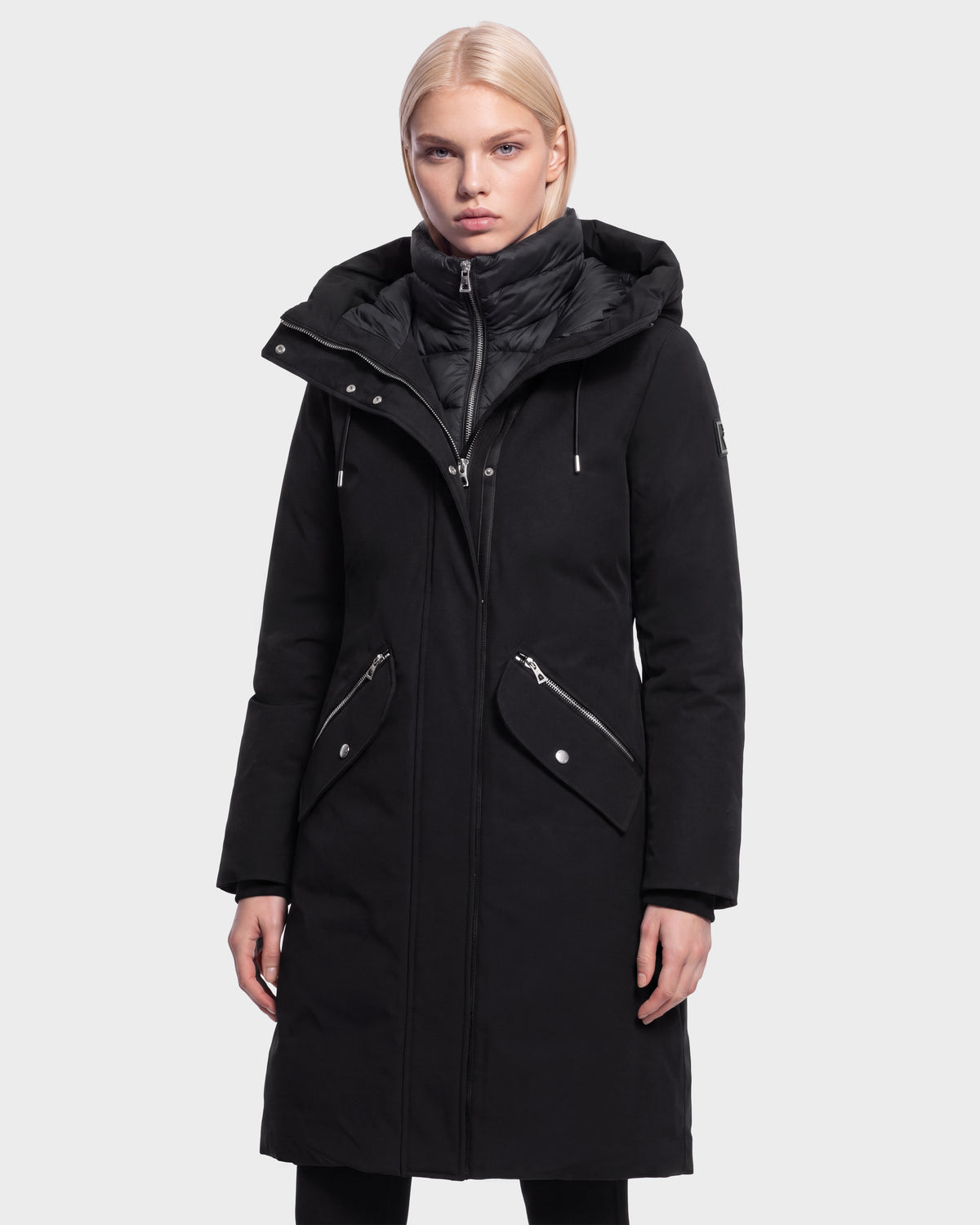A woman with blonde hair is wearing the Rudsak BRITTANY - 8125966 Black long winter coat, featuring a hood, zippered front, and snap pockets, while standing against a plain light background.