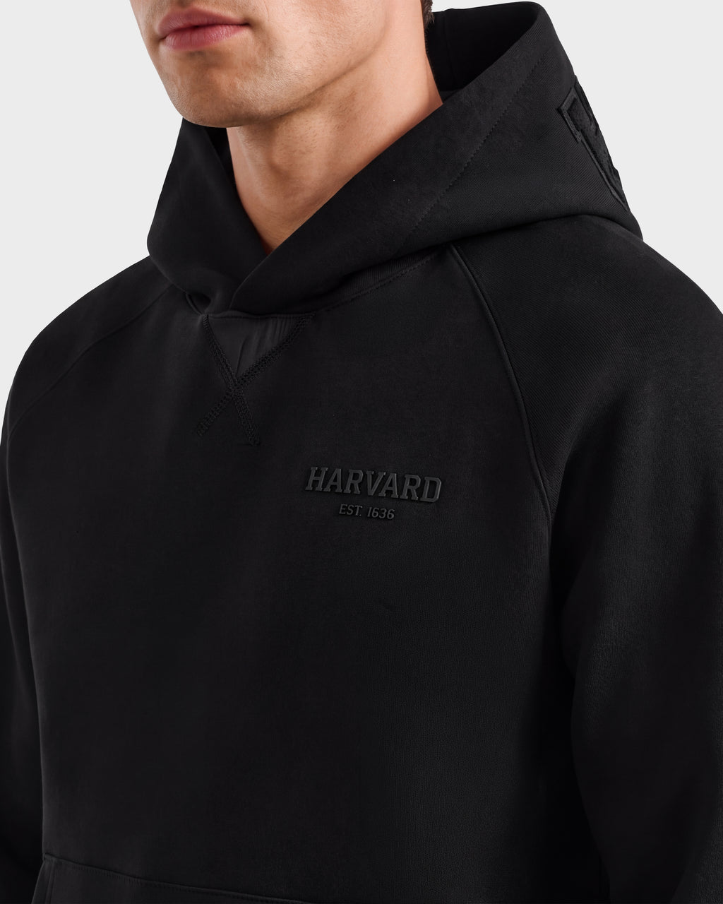 A person wearing the BARRY HV - H525500 Black hoodie by Rudsak, featuring the word HARVARD EST. 1636 elegantly embroidered on the chest. Only the lower half of their face is visible, lending an intriguing air of mystery reminiscent of Rudsak’s signature style.