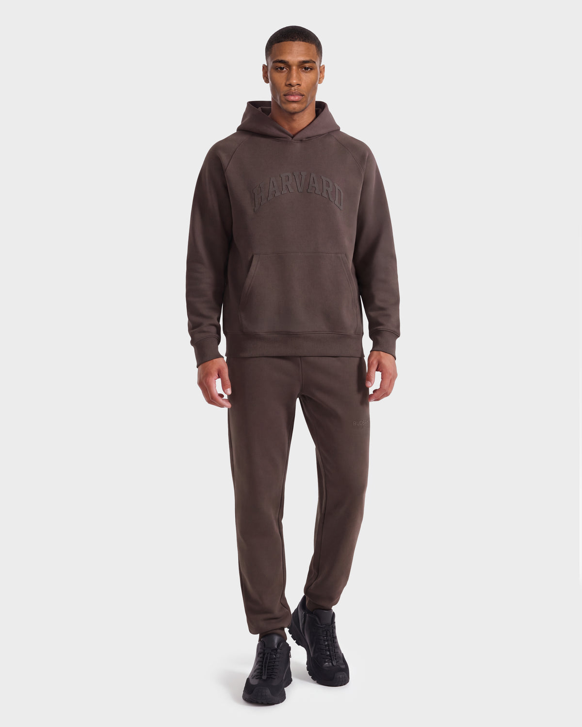 A man stands facing forward, wearing the limited-edition Rudsak BRIAR R HV - H525506 Espresso Harvard hoodie with matching sweatpants and black Rudsak sneakers, against a plain white background.