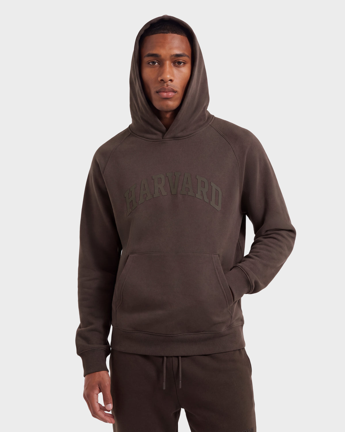 A man wearing the Rudsak BRIAR R HV - H525506 Espresso hoodie, a dark brown limited-edition piece with “Harvard” printed on the front, stands against a plain light background, looking at the camera with his hands at his sides.