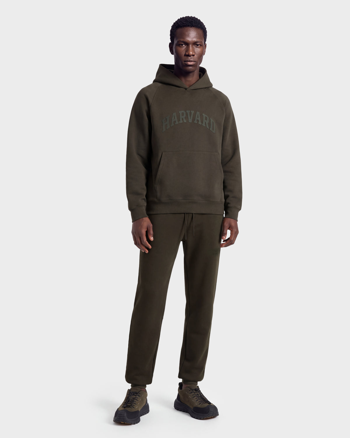 A man stands facing forward against a plain white background, wearing Rudsak’s limited-edition BRIAR R HV - H525506 Olive set: a matching dark green hoodie featuring “HARVARD” lettering and jogger pants, paired with dark sneakers.