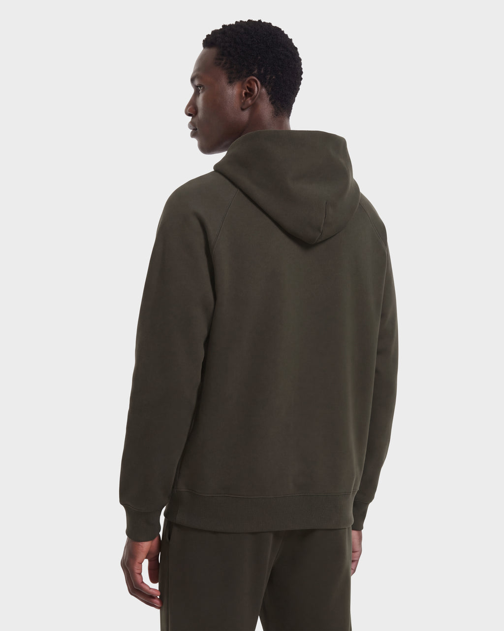 A person wearing the Rudsak BRIAR R HV - H525506 Olive limited-edition dark green hooded sweatshirt and matching pants stands facing away, looking to the side, against a plain white background.