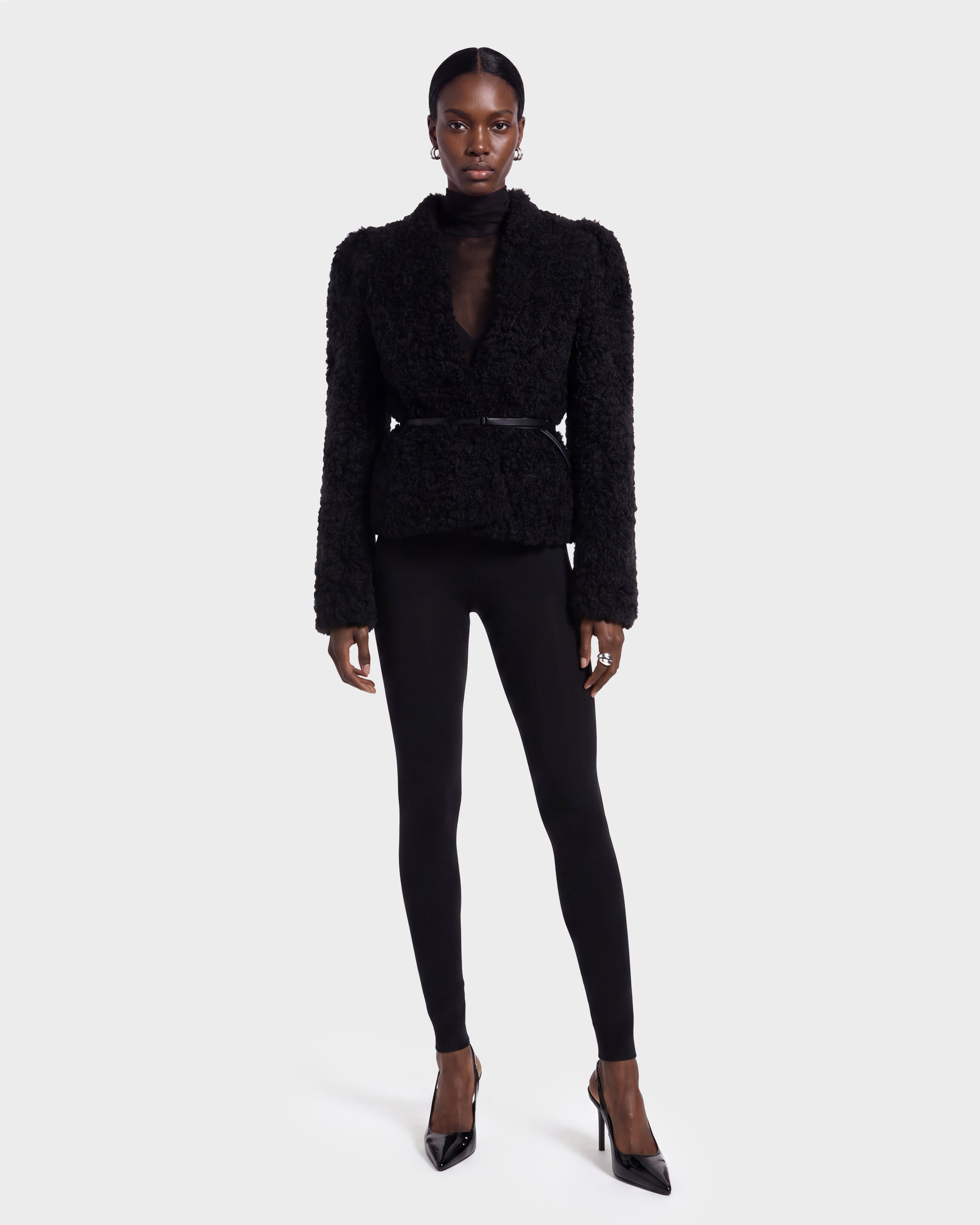 A woman wearing the BRINLEY - 8126980 Black textured jacket by Rudsak, a sheer black top, black leggings, and black high heels stands against a plain light gray background.