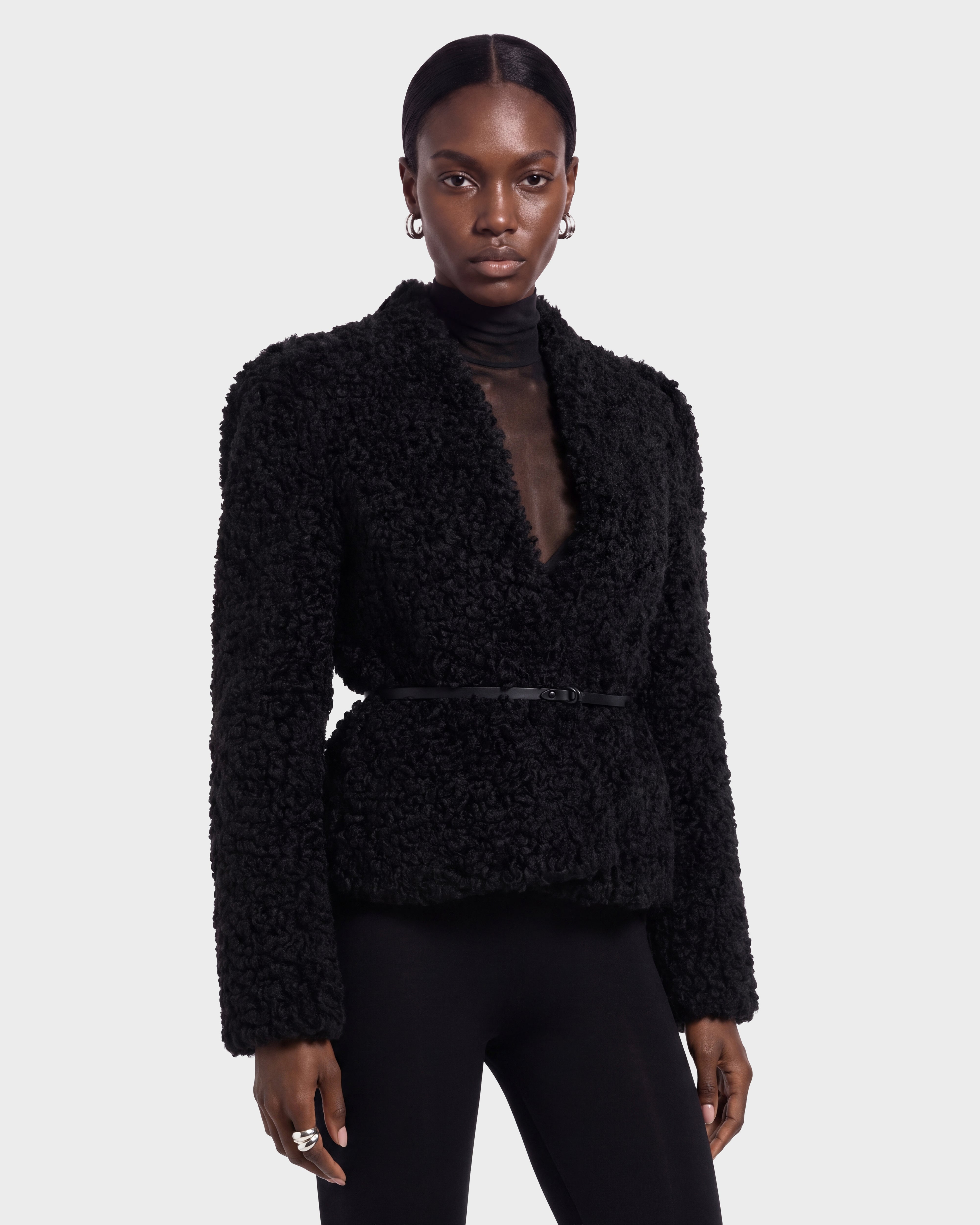 A person wearing the BRINLEY - 8126980 Black jacket by Rudsak, featuring a textured design with a belt, stands facing forward against a plain light background, paired with a sheer black top and black pants.