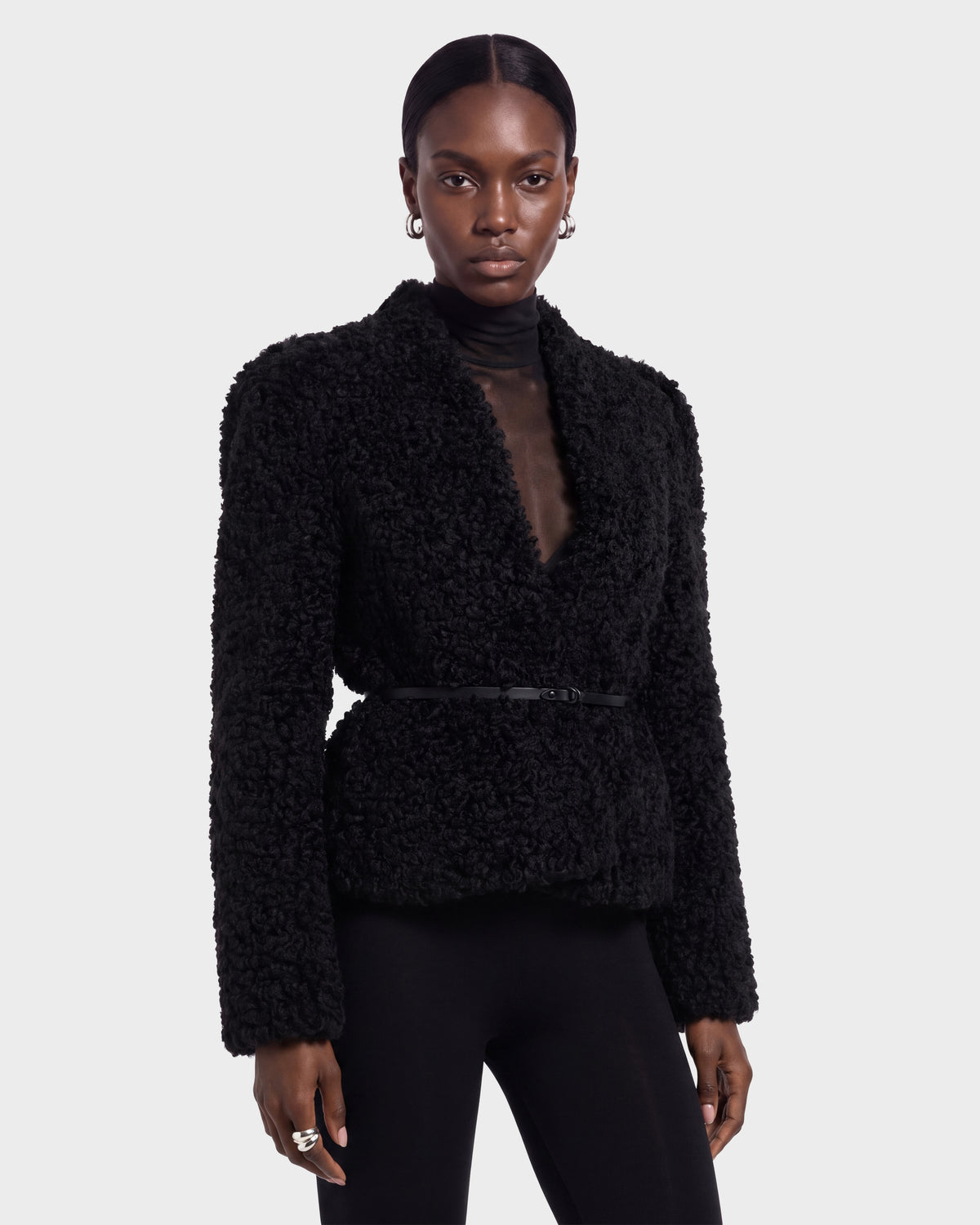 A person wearing the BRINLEY - 8126980 Black jacket by Rudsak, featuring a textured design with a belt, stands facing forward against a plain light background, paired with a sheer black top and black pants.
