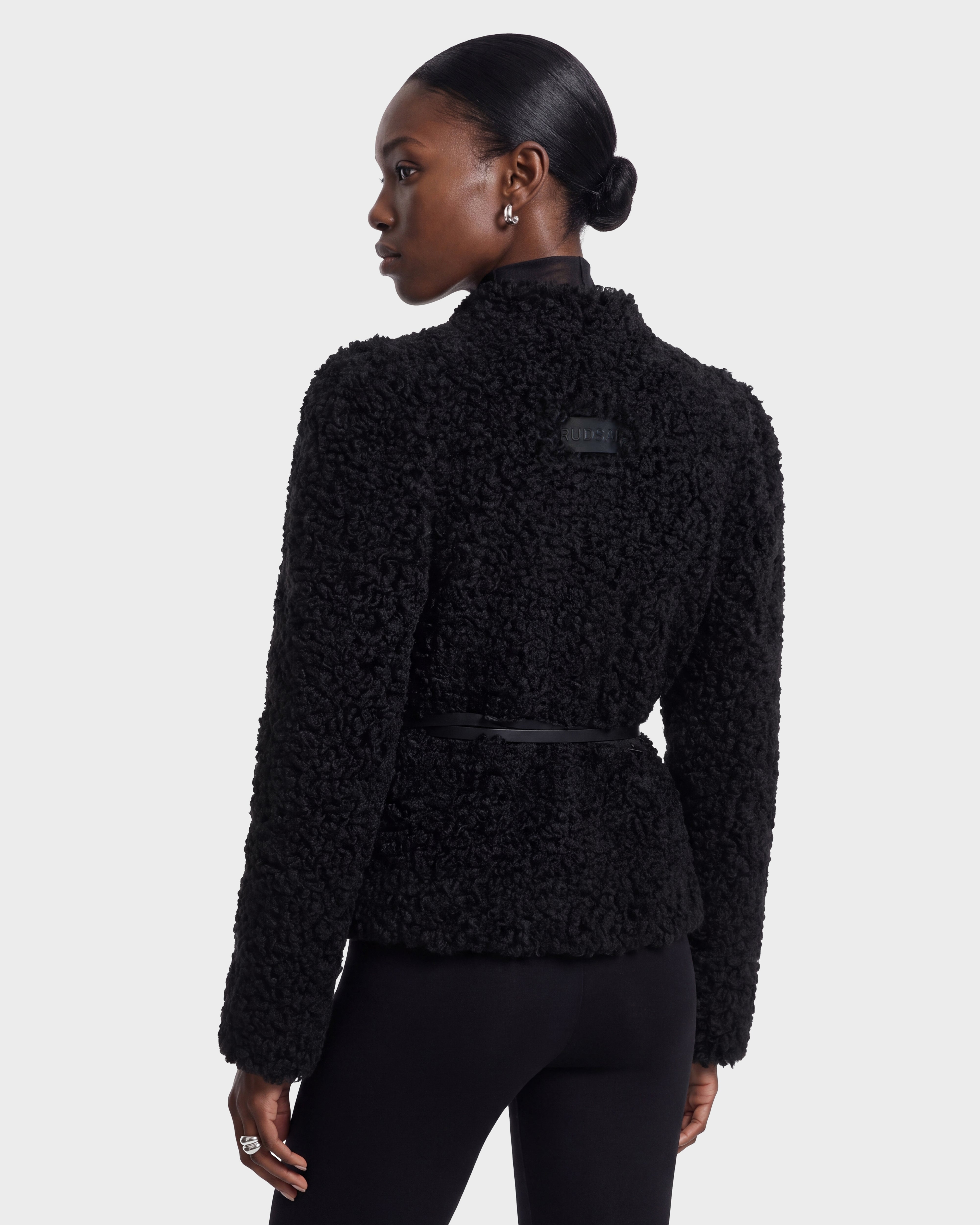 A woman is seen from the back wearing the BRINLEY - 8126980 Black jacket by Rudsak, featuring a textured design and a black belt. Her dark hair is styled in a neat bun, and she pairs the jacket with black pants.