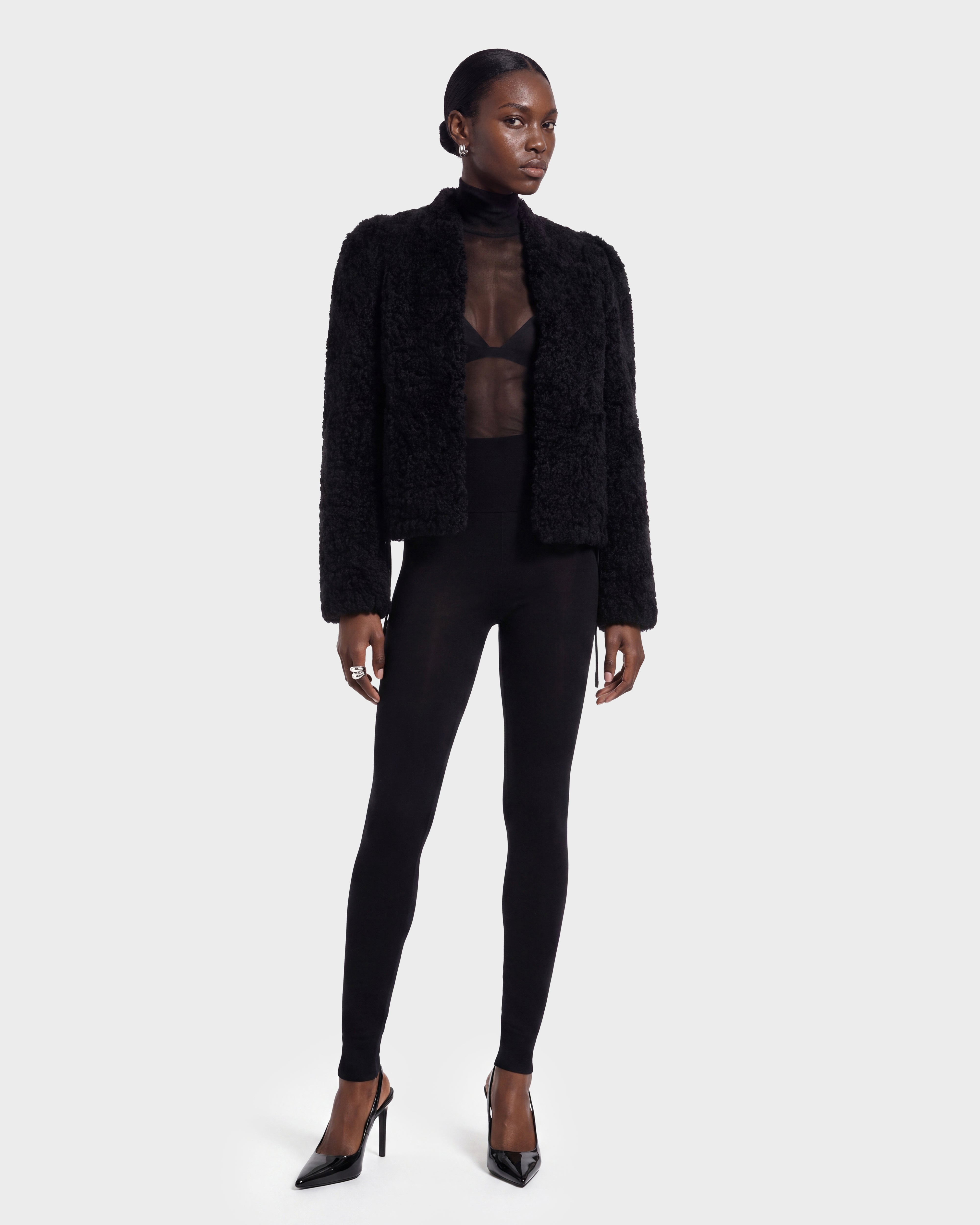 A woman stands against a plain background, wearing the BRINLEY - 8126980 Black jacket by Rudsak paired with a sheer black top, black bra, fitted black leggings, and black high-heeled shoes. Her hair is pulled back and her expression is neutral.