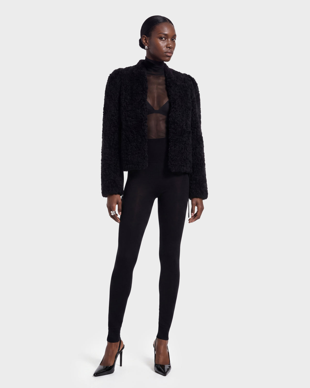 A woman stands against a plain background, wearing the BRINLEY - 8126980 Black jacket by Rudsak paired with a sheer black top, black bra, fitted black leggings, and black high-heeled shoes. Her hair is pulled back and her expression is neutral.
