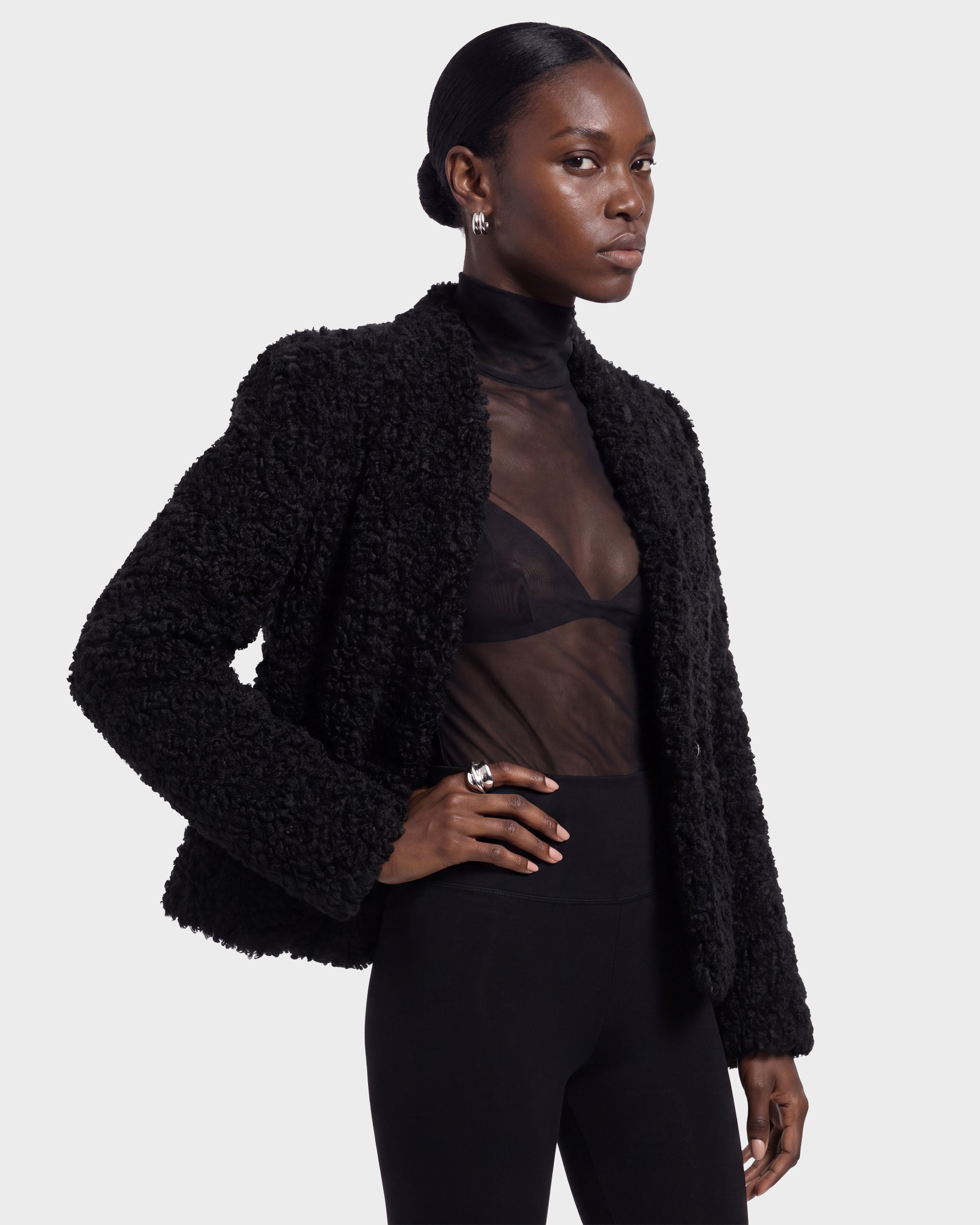 A woman models the BRINLEY - 8126980 Black textured jacket by Rudsak over a sheer black top and black pants. She wears her hair pulled back, accessorizing with silver hoop earrings and a ring, standing against a plain light background.