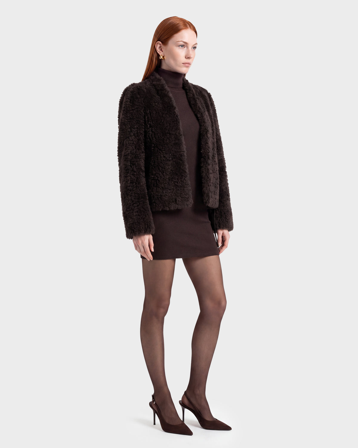 A woman with long red hair wears the Rudsak BRINLEY - 8126980 Espresso dark faux fur jacket over a brown turtleneck dress, paired with sheer black tights and dark pointed heels, standing against a plain light background.