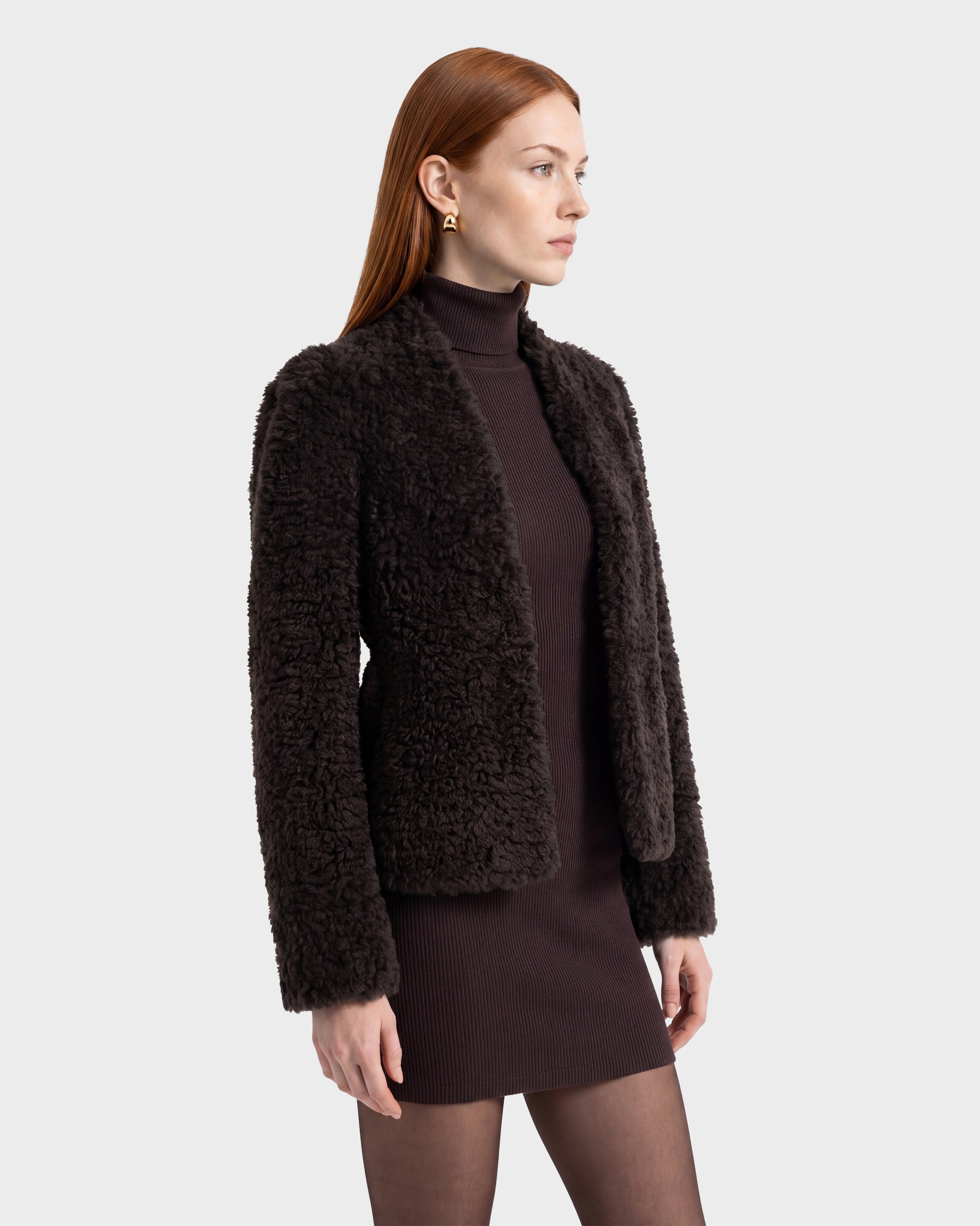 A woman with long red hair wears the Rudsak BRINLEY - 8126980 Espresso dark curly faux fur jacket over a ribbed dark dress, black tights, and gold earrings, standing against a light gray background, facing slightly to the right.