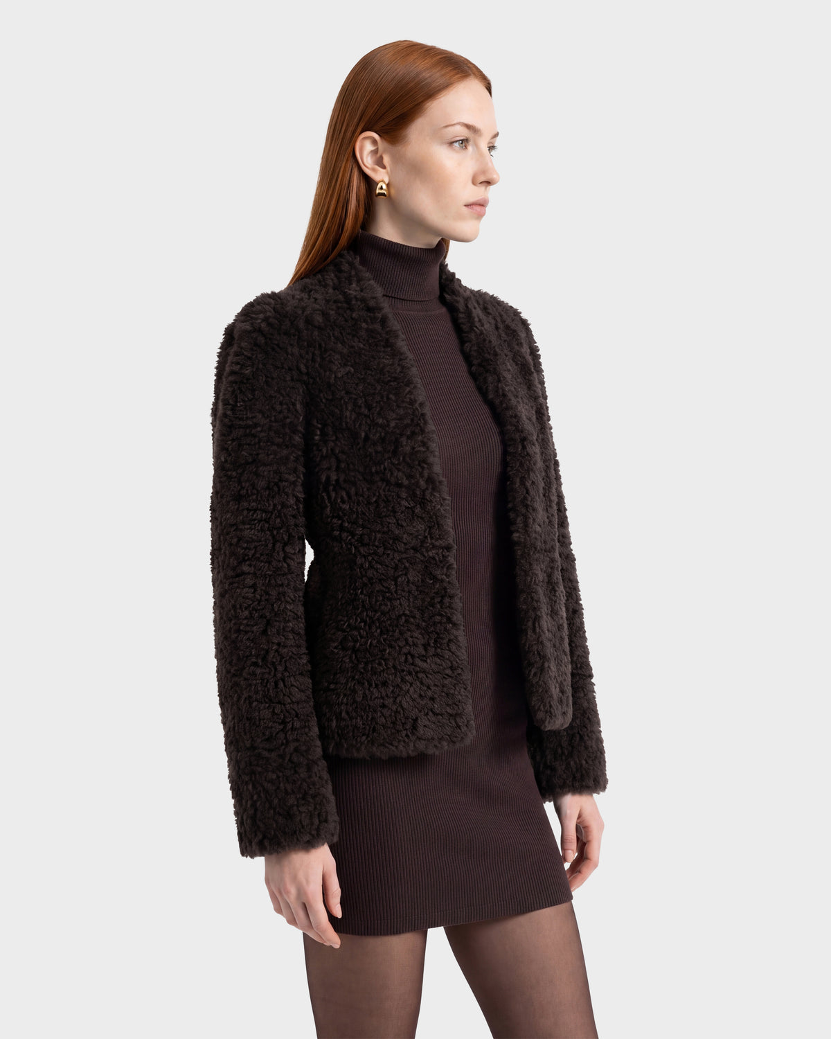 A woman with long red hair wears the Rudsak BRINLEY - 8126980 Espresso dark curly faux fur jacket over a ribbed dark dress, black tights, and gold earrings, standing against a light gray background, facing slightly to the right.