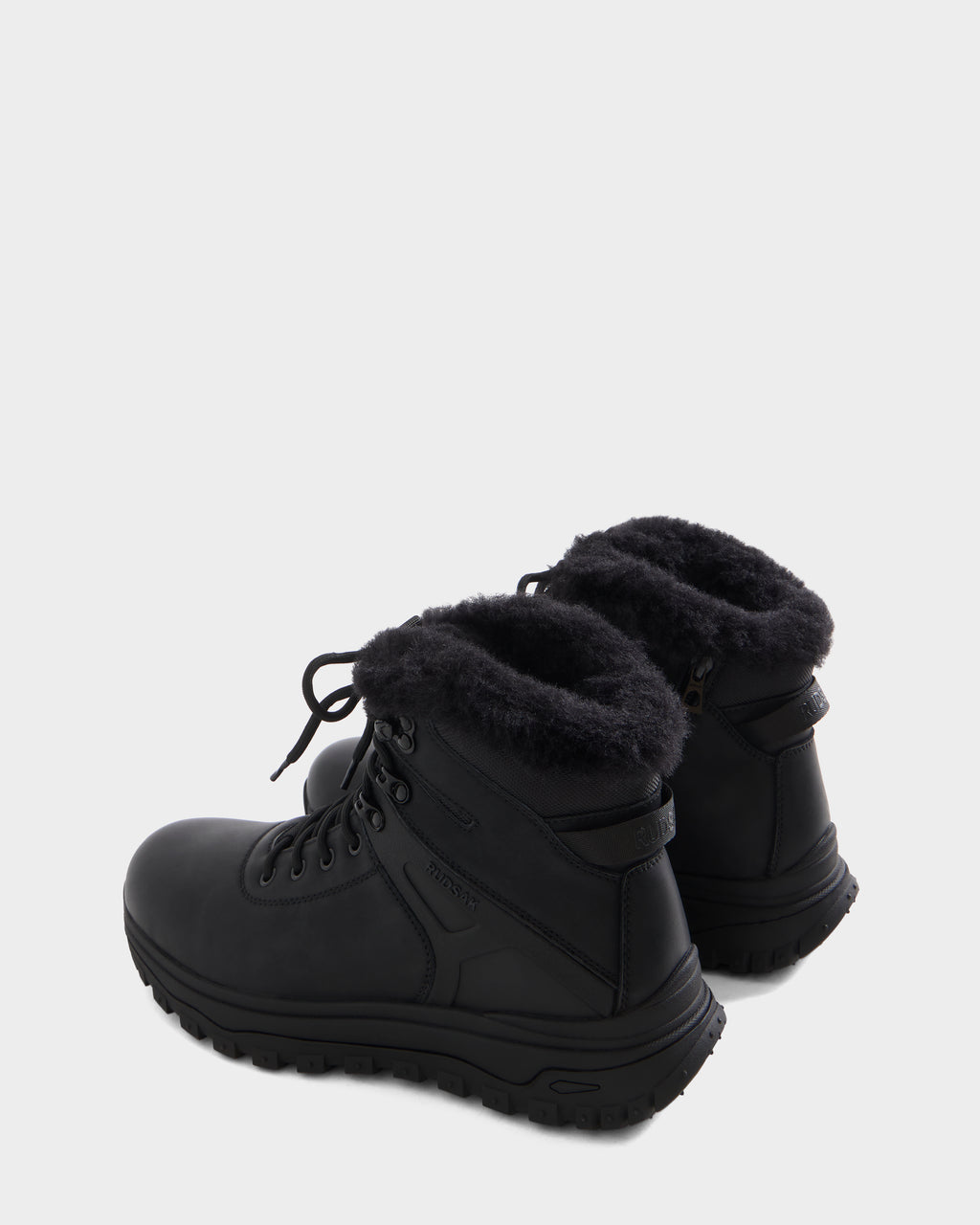 The BRYSON - 6226504 Black by Rudsak are black winter boots with thick rubber soles and black faux fur ankle lining, featuring a lugged outsole, shown from a rear three-quarter angle on a plain light background.