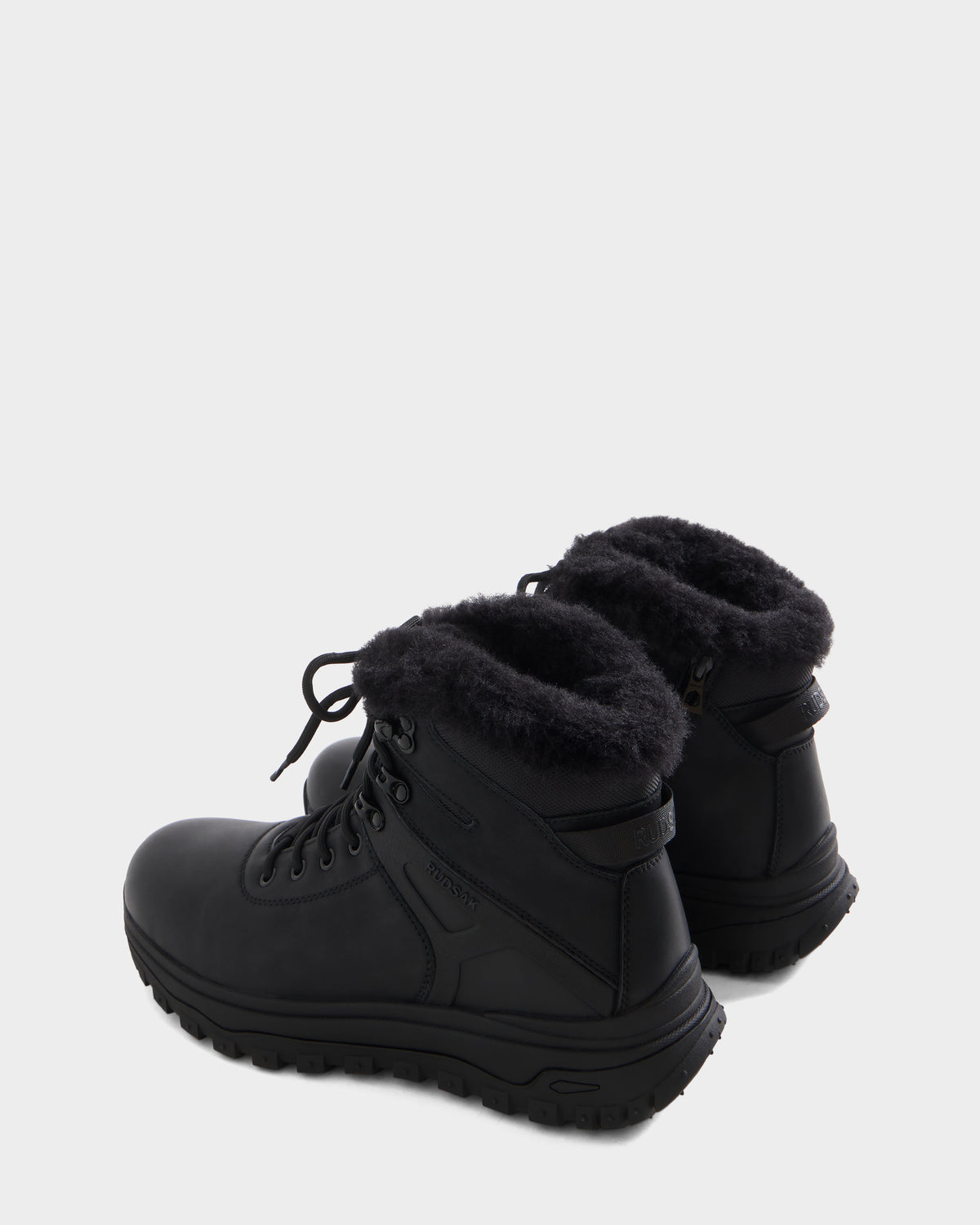 The BRYSON - 6226504 Black by Rudsak are black winter boots with thick rubber soles and black faux fur ankle lining, featuring a lugged outsole, shown from a rear three-quarter angle on a plain light background.
