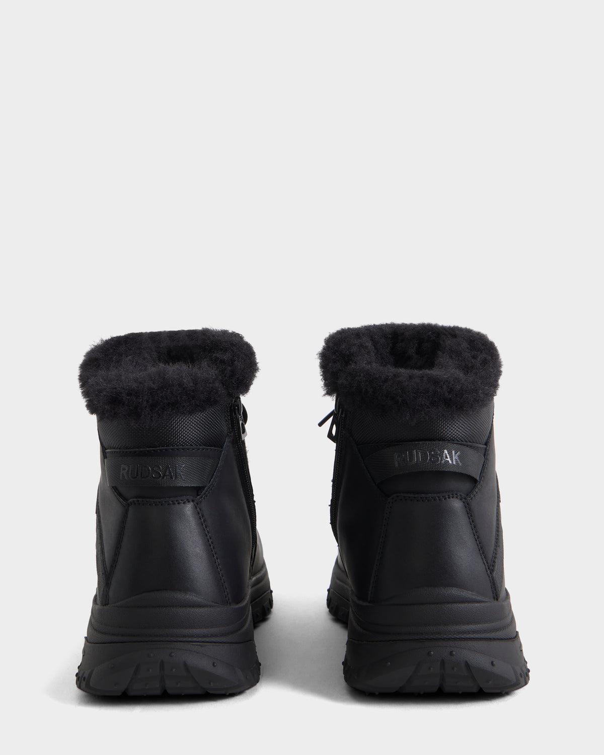 A pair of BRYSON - 6226504 Black winter boots with fur trim and lugged outsole, seen from the back on a white background. The Rudsak logo appears on the heel of each boot.