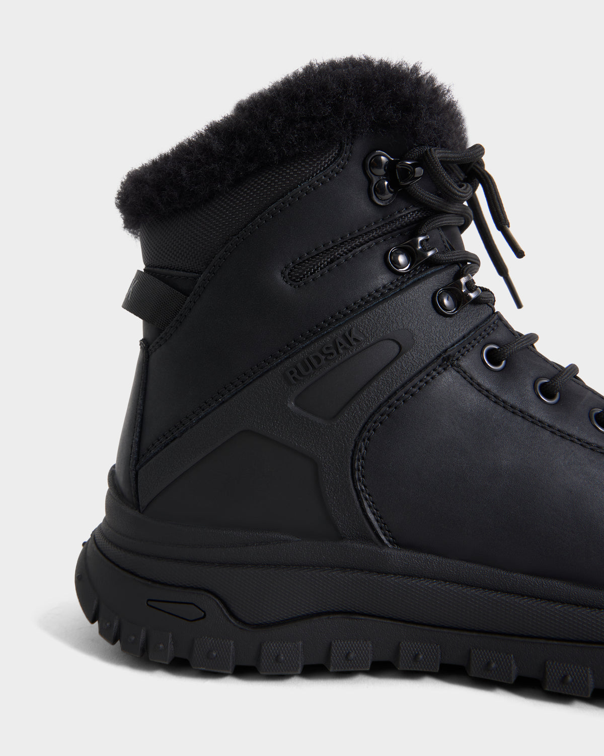 A close-up of the BRYSON - 6226504 Black boot shows a black lace-up design with metal eyelets, a rugged lugged sole, fluffy collar, and Rudsak embossed on the side, photographed on a white background.