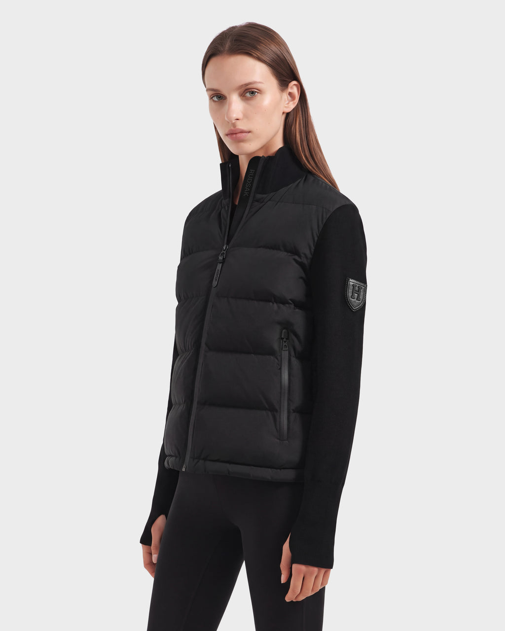 A person wearing the Rudsak CAMILLE HV - H125500 Black jacket styled as a quilted puffer vest over a black long-sleeve top and black pants stands against a plain, light background, looking toward the camera with a neutral expression.