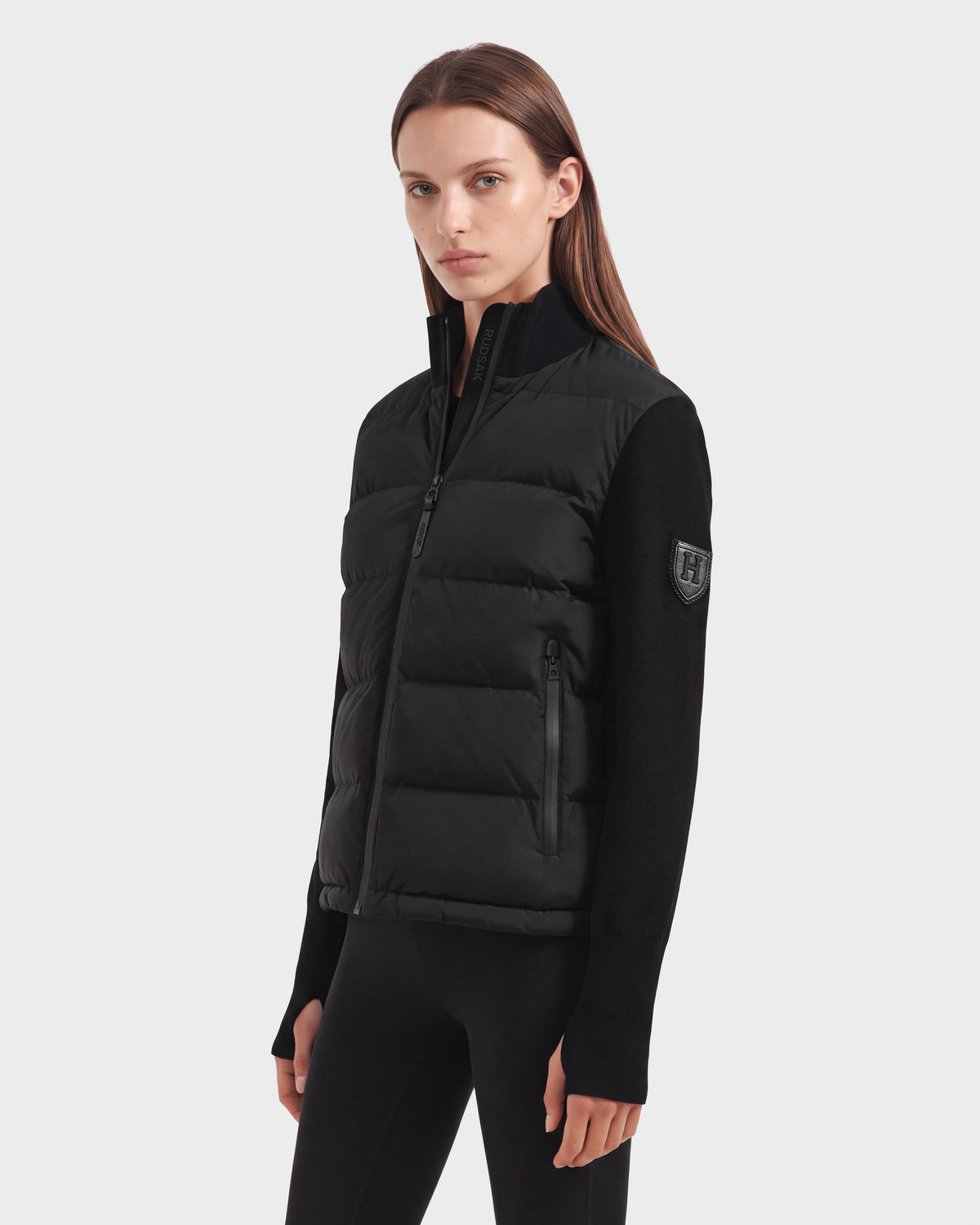 A person wearing the Rudsak CAMILLE HV - H125500 Black jacket styled as a quilted puffer vest over a black long-sleeve top and black pants stands against a plain, light background, looking toward the camera with a neutral expression.