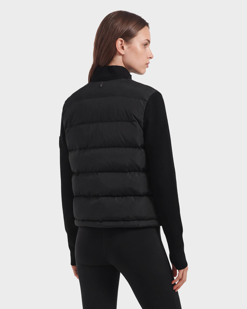 Woman with straight brown hair wearing the limited-edition Rudsak CAMILLE HV - H125500 Black down jacket over a black long-sleeve top and black leggings, standing and facing away, looking to the left against a plain light background.
