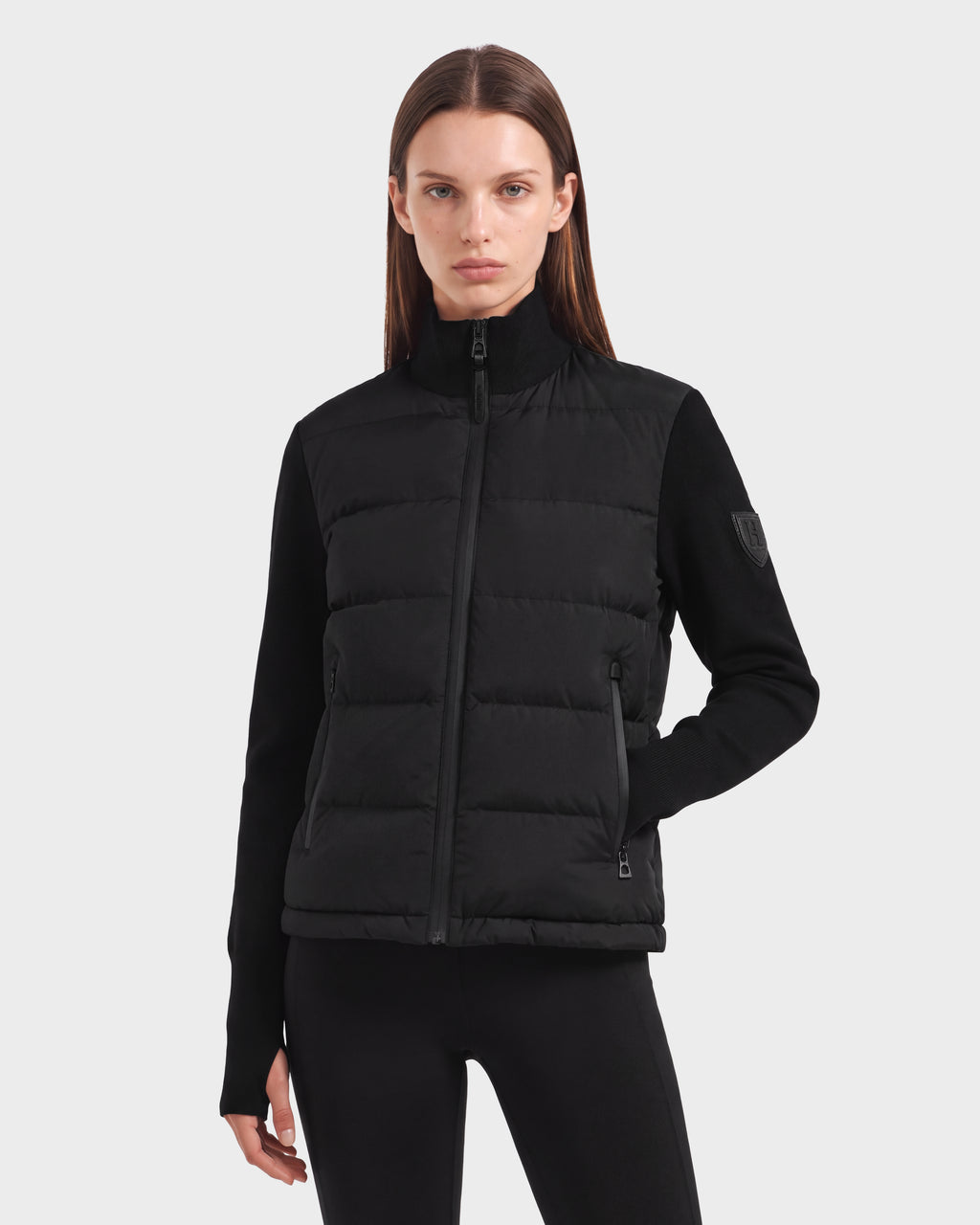 A woman with straight brown hair wears the Rudsak CAMILLE HV - H125500 Black limited-edition down jacket and black pants, standing against a plain light background with a neutral expression.