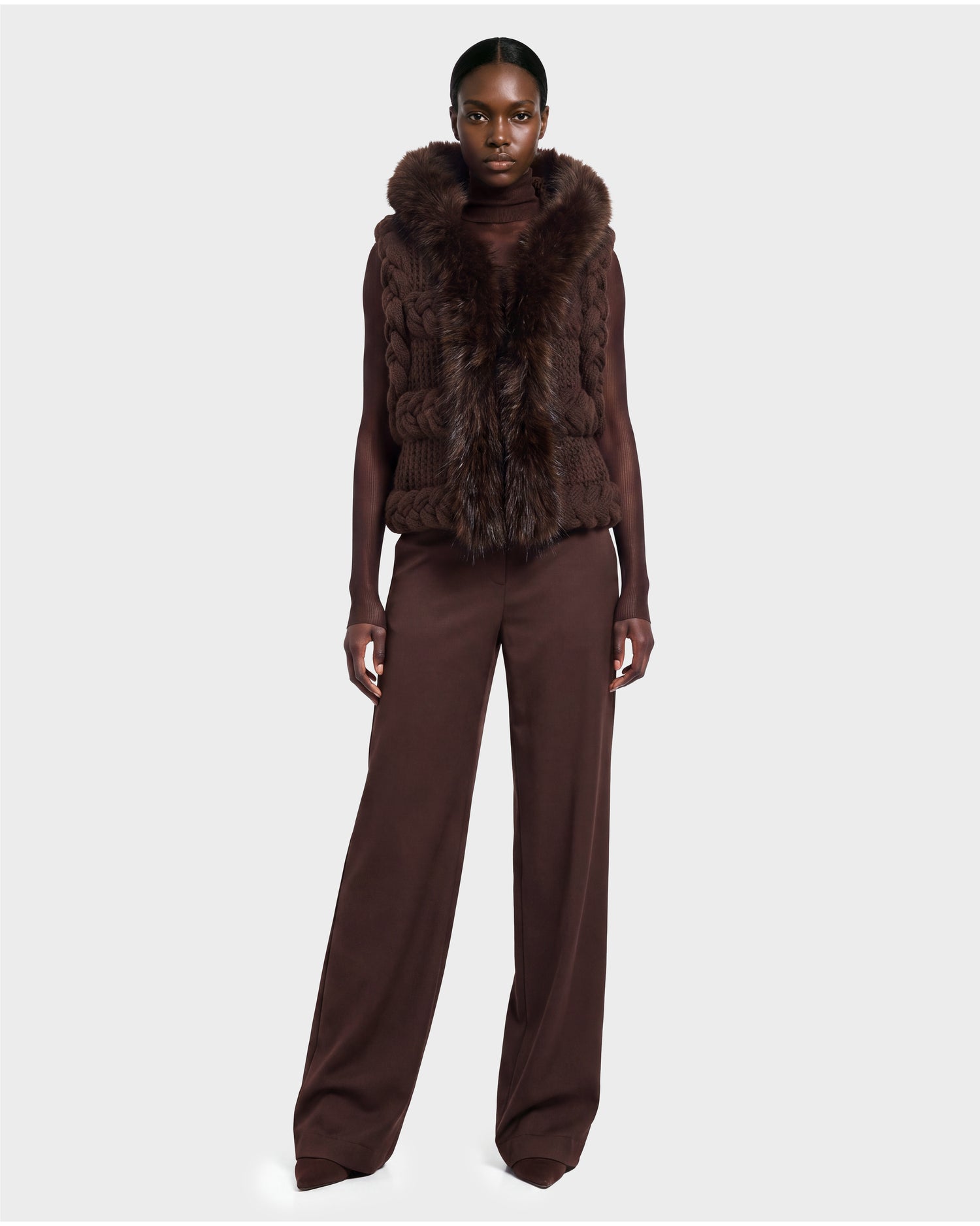 A woman stands facing forward, wearing the Rudsak CICELY - 8526686 Espresso vest—an elegant dark brown fur-trimmed piece—layered over a long-sleeve top and coordinating wide-leg trousers in rich espresso hues. Set against a plain light gray background, this stylish ensemble is available for pre-order and will ship on September 15th.