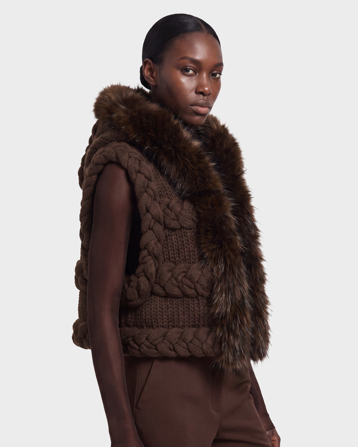 A woman wears the CICELY - 8526686 Espresso dark brown knitted vest by Rudsak, featuring thick braided details and faux fur trim, layered over a long-sleeved brown top and brown pants against a plain light background. This stylish piece is available for pre-order now and will ship on September 15th.