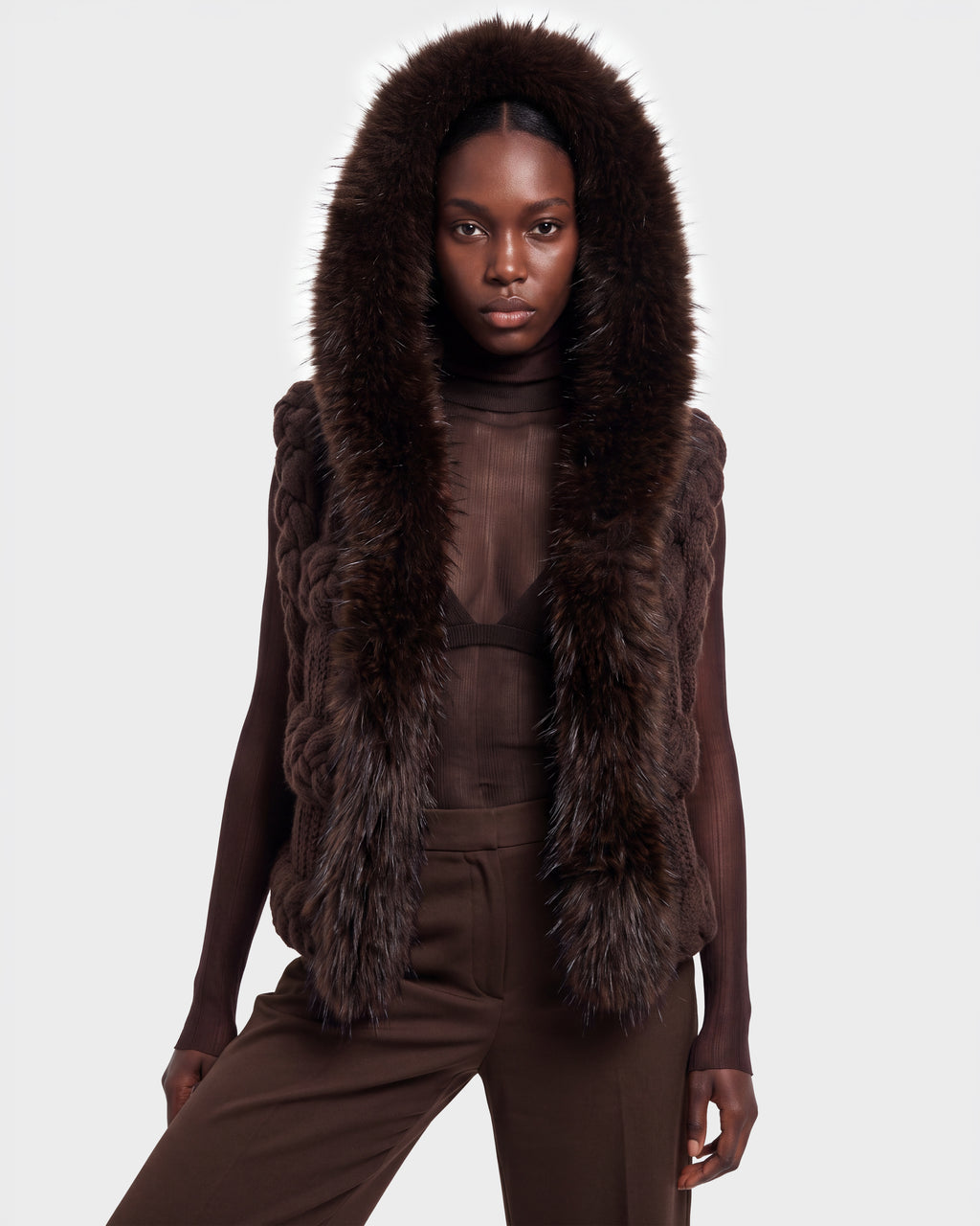 A woman stands facing forward against a plain light background, dressed in the CICELY - 8526686 Espresso by Rudsak—a sheer brown turtleneck top and matching pants, layered with a sleeveless, fur-trimmed hooded vest. This stylish ensemble is now available for pre-order and will ship on September 15th.