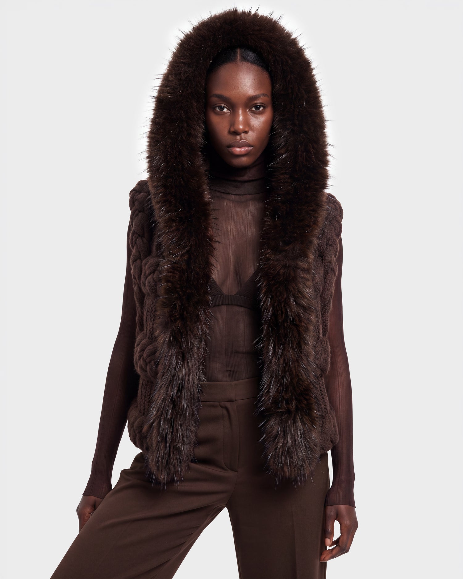 A woman stands facing forward against a plain light background, dressed in the CICELY - 8526686 Espresso by Rudsak—a sheer brown turtleneck top and matching pants, layered with a sleeveless, fur-trimmed hooded vest. This stylish ensemble is now available for pre-order and will ship on September 15th.