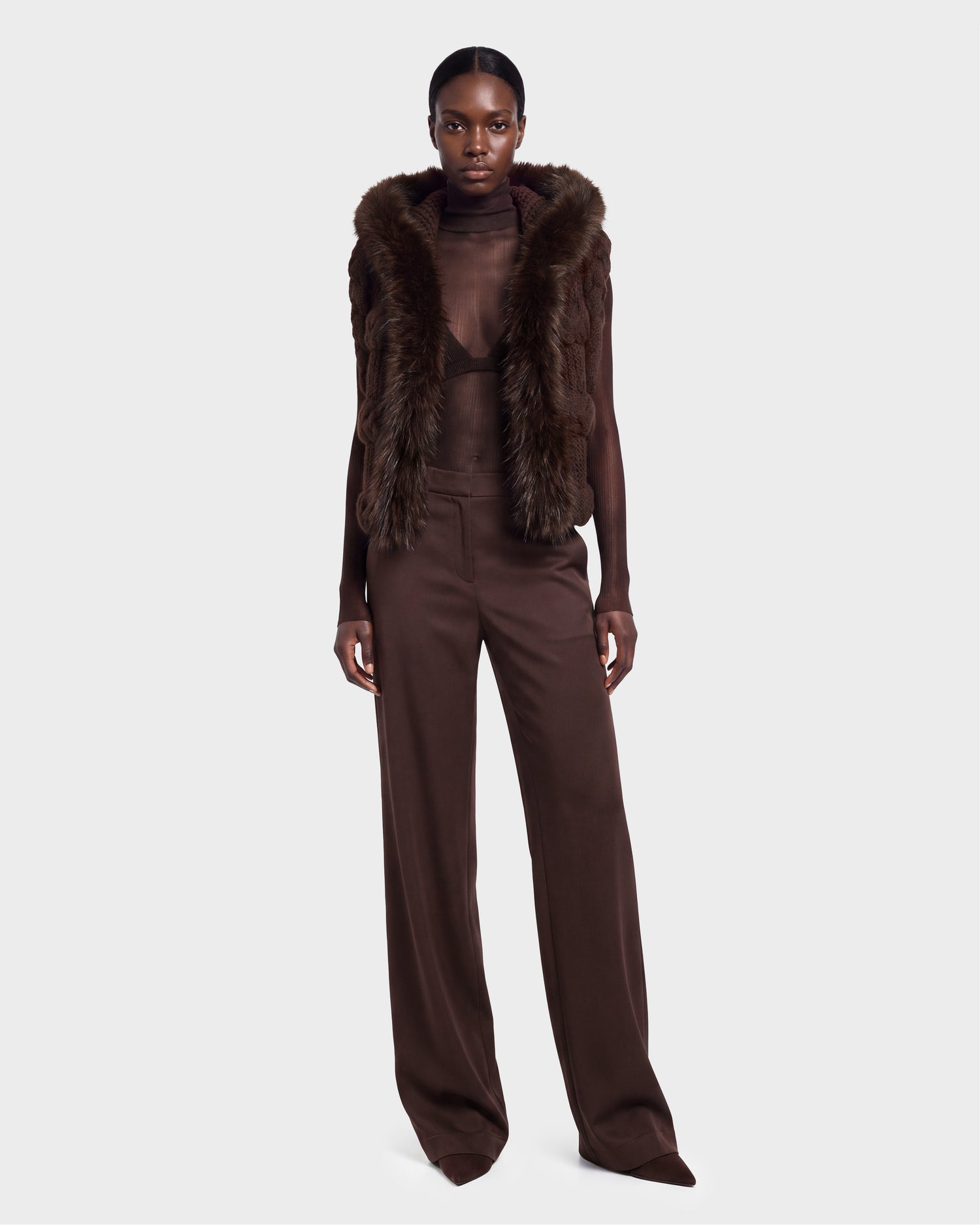 A person stands facing forward, wearing the CICELY - 8526686 Espresso sleeveless faux fur vest by Rudsak over a sheer brown top and wide-leg brown pants, against a plain light background. This stylish outfit is available for pre-order and will ship September 15th.