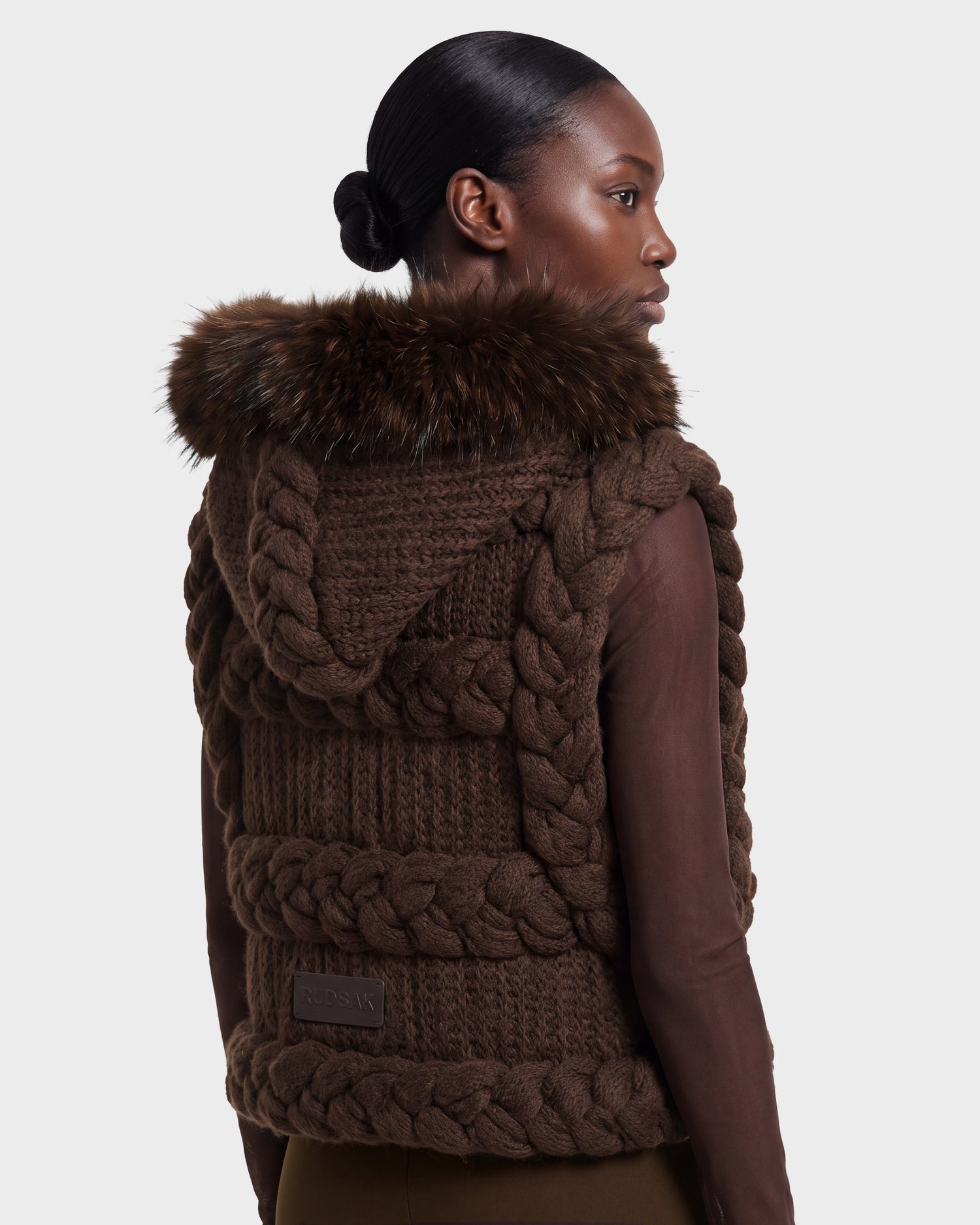 A woman with smooth, pulled-back hair wears the Rudsak CICELY - 8526686 Espresso: a brown, chunky knit vest with braided details and a fur-trimmed hood, layered over a brown long-sleeve top as she faces slightly to the right. This stylish piece is available for pre-order and will ship September 15th.