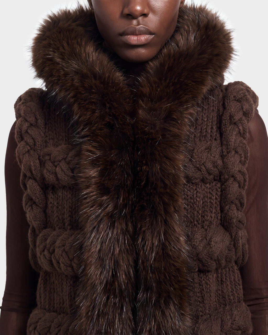 A person wearing the CICELY - 8526686 Espresso by Rudsak—a chunky, dark brown knit vest featuring large braided patterns and a thick, fluffy brown fur collar—shown from shoulders to chin. Available for pre-order now and set to ship September 15th.