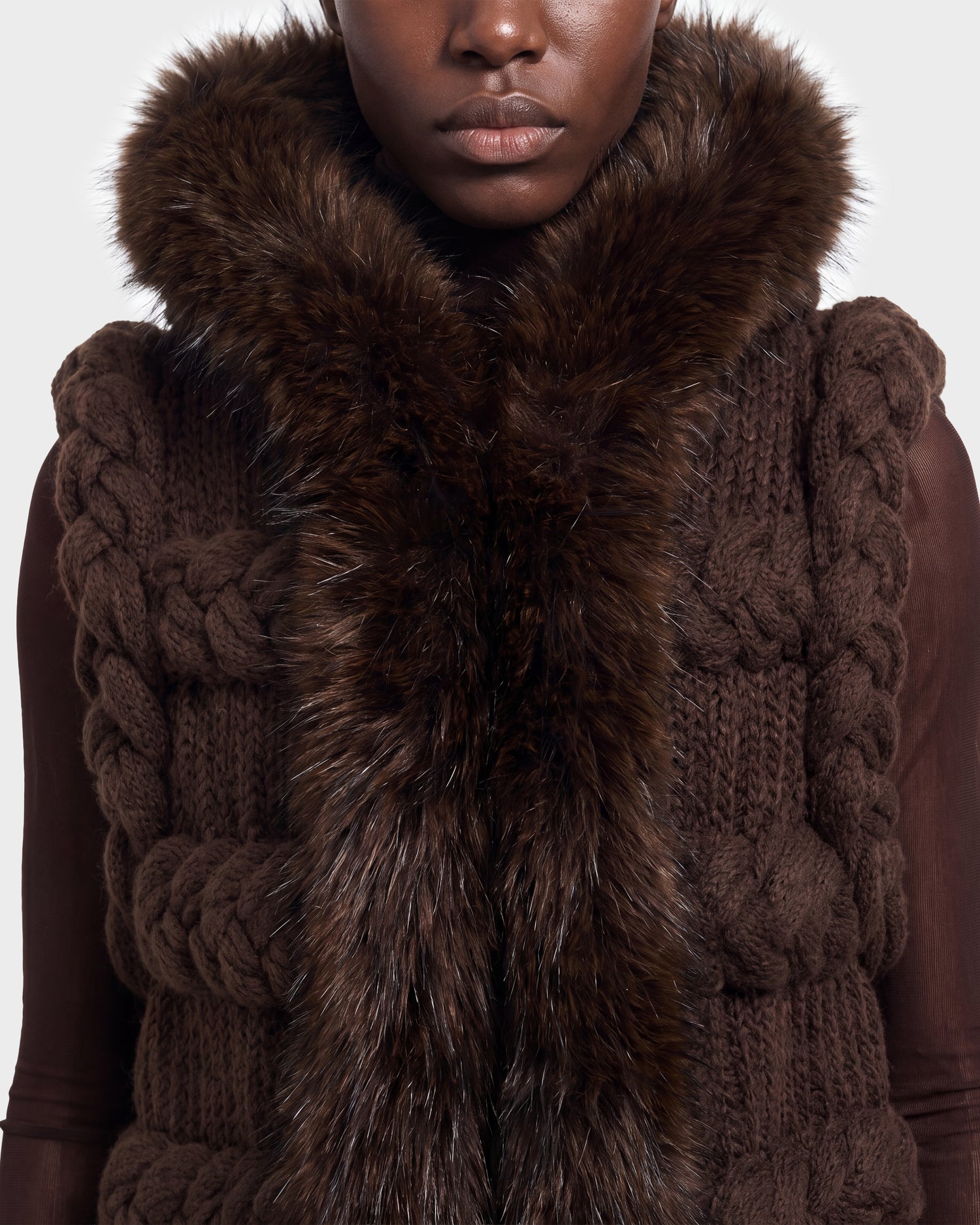 A person wearing the CICELY - 8526686 Espresso by Rudsak—a chunky, dark brown knit vest featuring large braided patterns and a thick, fluffy brown fur collar—shown from shoulders to chin. Available for pre-order now and set to ship September 15th.