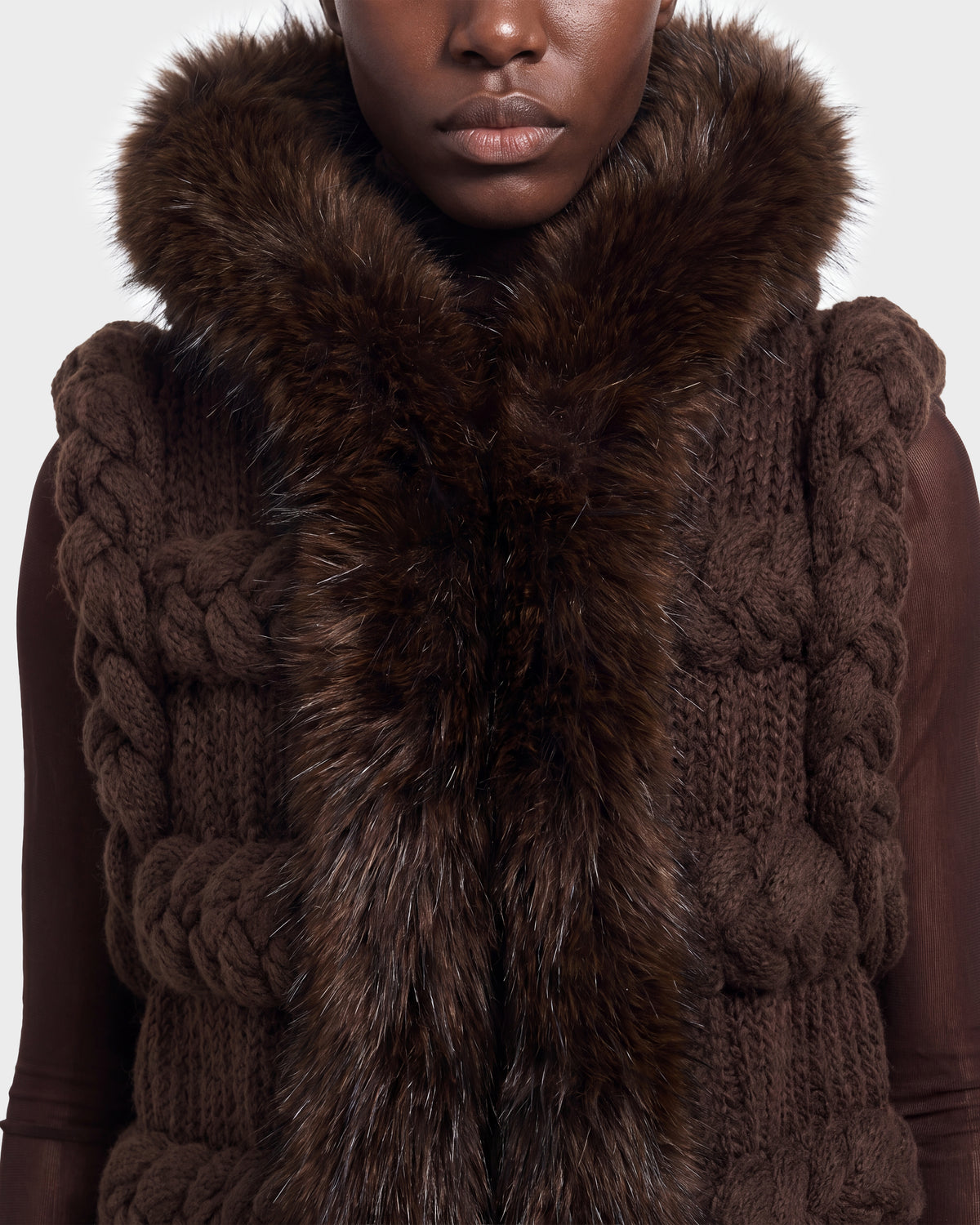 A person wearing the CICELY - 8526686 Espresso by Rudsak—a chunky, dark brown knit vest featuring large braided patterns and a thick, fluffy brown fur collar—shown from shoulders to chin. Available for pre-order now and set to ship September 15th.
