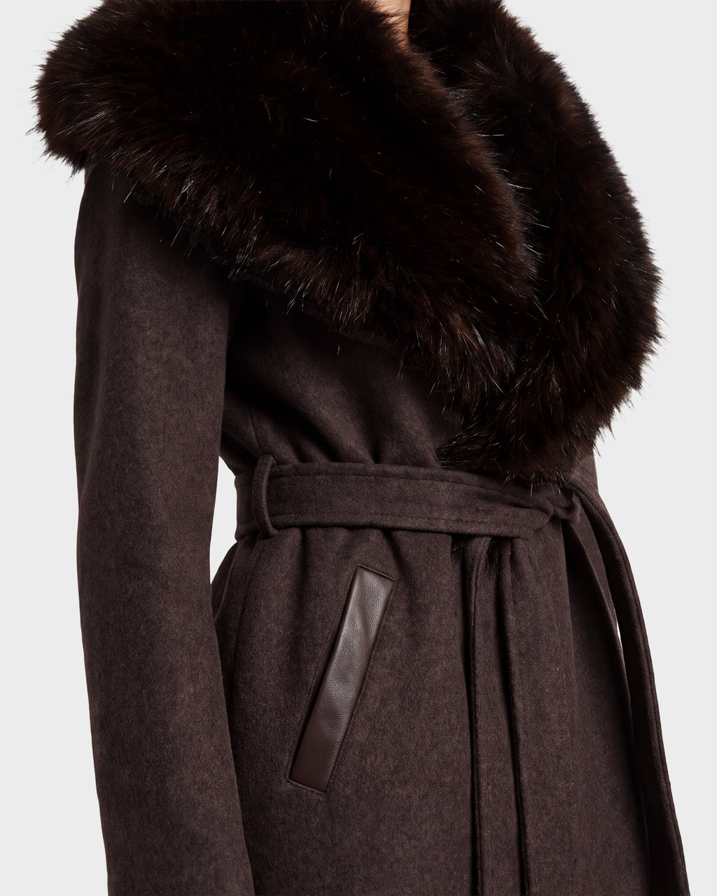 A close-up of a person wearing the Rudsak CINDY - 8126526 Heather Cacao coat, featuring dark brown wool, a large fluffy faux fur collar, leather-trimmed pockets, and a stylish belt.