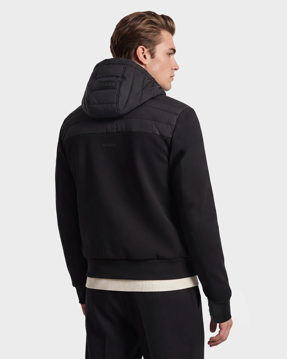 A man with light brown hair is shown from the back wearing the Rudsak DEE MAT - 6124028 Black urban jacket, featuring a hood and ribbed hem and cuffs, paired with black pants. The background is plain light gray.