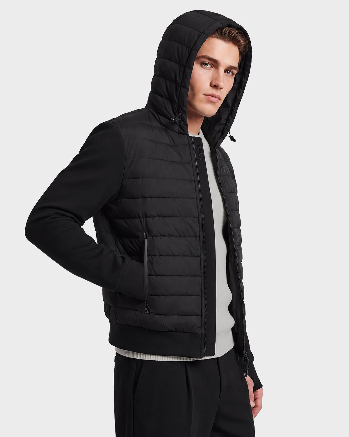 A man wearing the DEE MAT - 6124028 Black Rudsak men's lightweight hybrid down jacket over a light-colored shirt stands with one hand in his pocket, looking slightly to the side. The background is plain and light-colored.