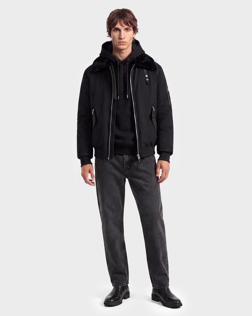 A man stands facing forward, wearing a modern outerwear look featuring the Rudsak DYLAN - 6125593 Black men’s aviator bomber jacket with a faux fur collar, paired with a black hoodie, dark jeans, and black shoes against a plain light gray background.