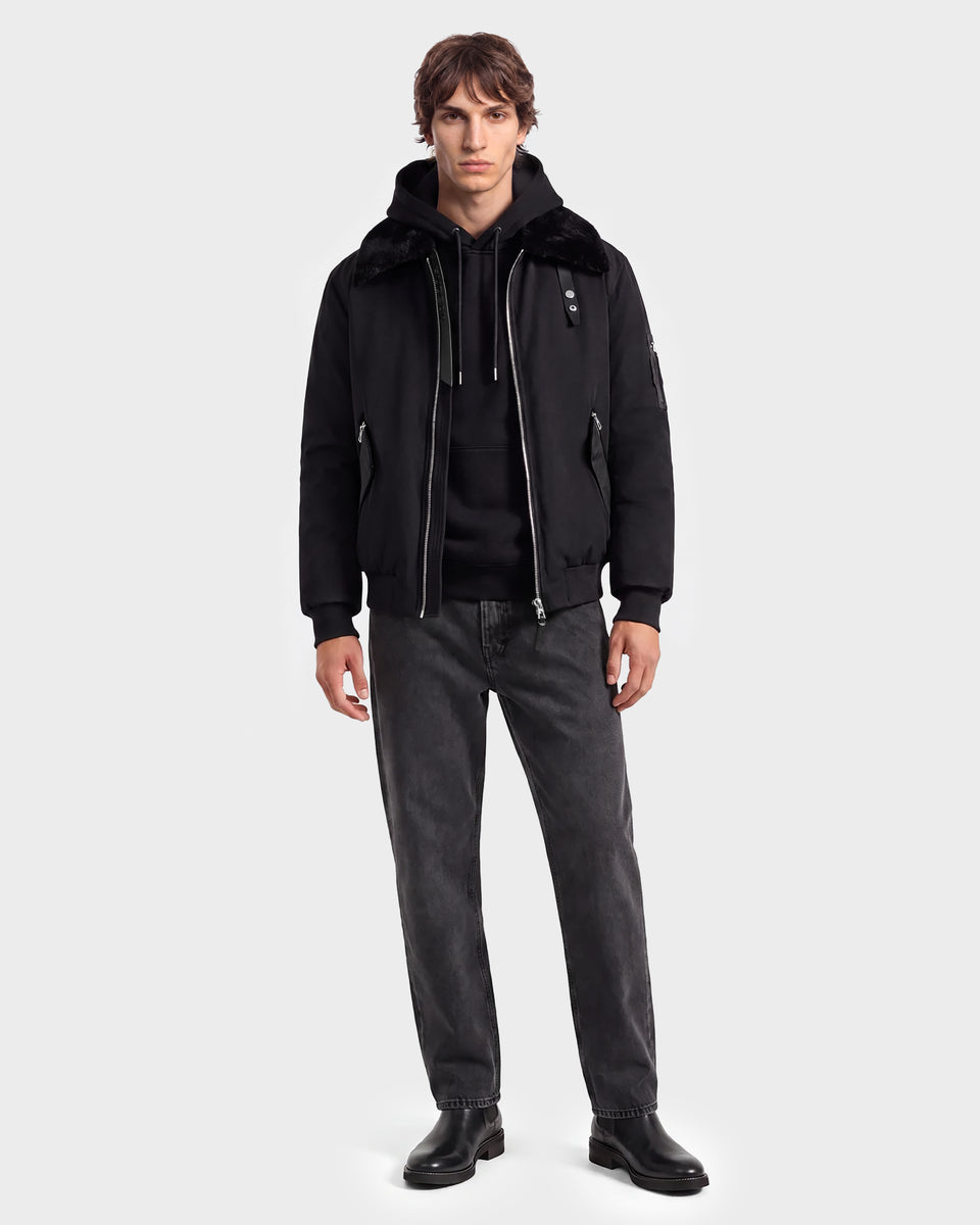 A man stands facing forward, wearing a modern outerwear look featuring the Rudsak DYLAN - 6125593 Black men’s aviator bomber jacket with a faux fur collar, paired with a black hoodie, dark jeans, and black shoes against a plain light gray background.