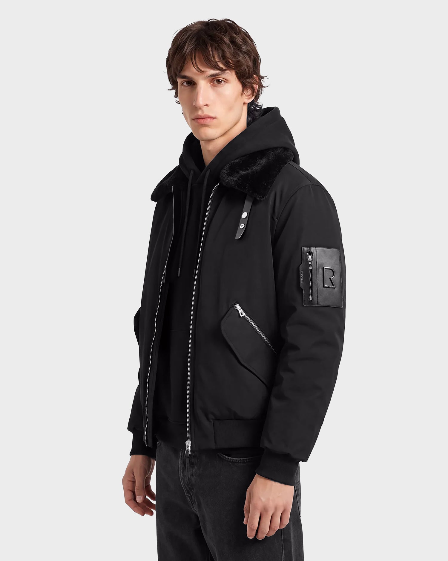 A young person with short brown hair is standing against a plain light background, modeling the Rudsak DYLAN - 6125593 Black men’s aviator bomber jacket. The jacket features a faux fur collar, zippered pockets, and an "R" patch on the sleeve, highlighting its sleek modern outerwear style.
