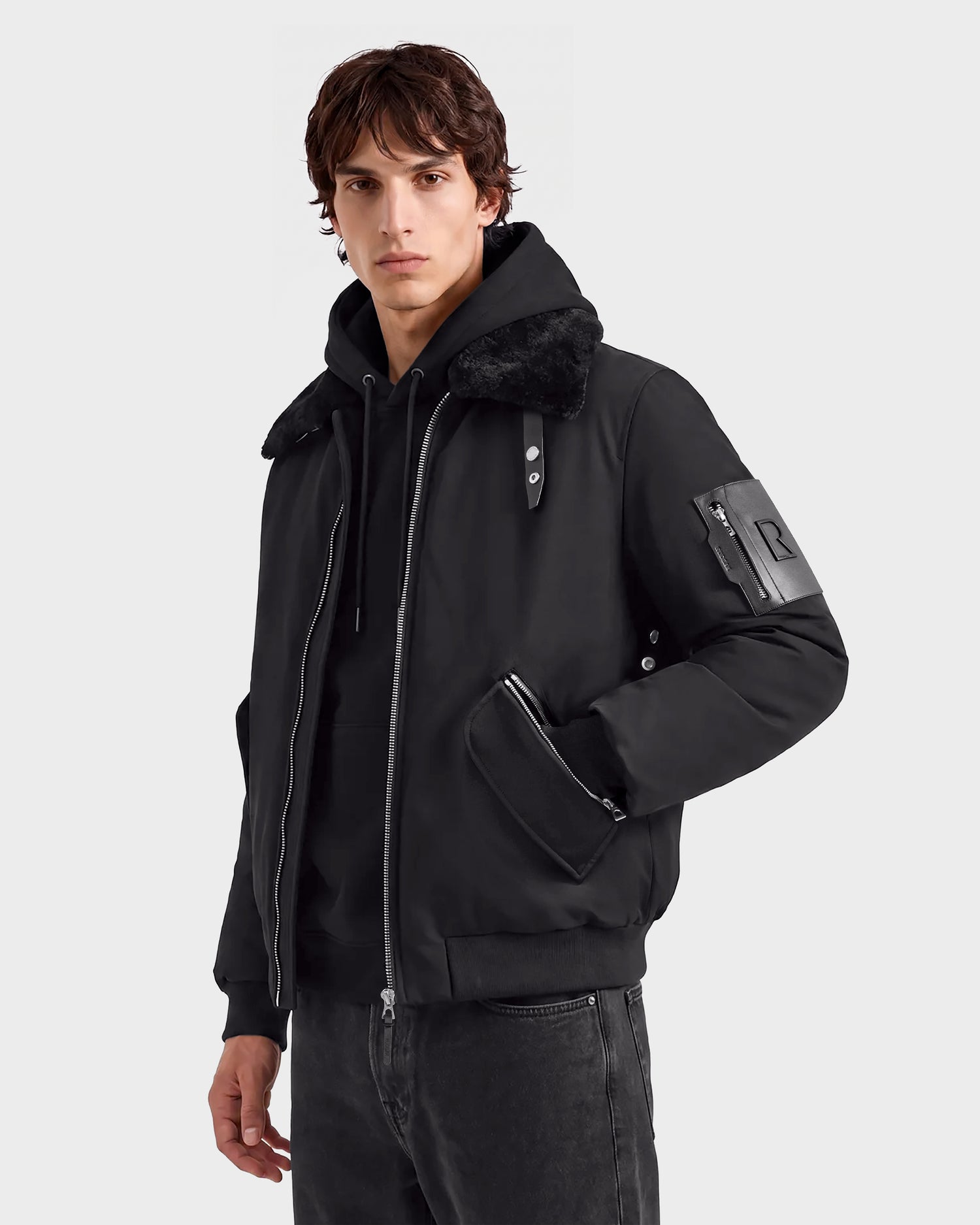 A person wearing the Rudsak DYLAN - 6125593 Black men’s aviator bomber jacket with a faux fur collar, paired with black jeans and a serious expression, stands against a plain light gray background, highlighting modern outerwear style.