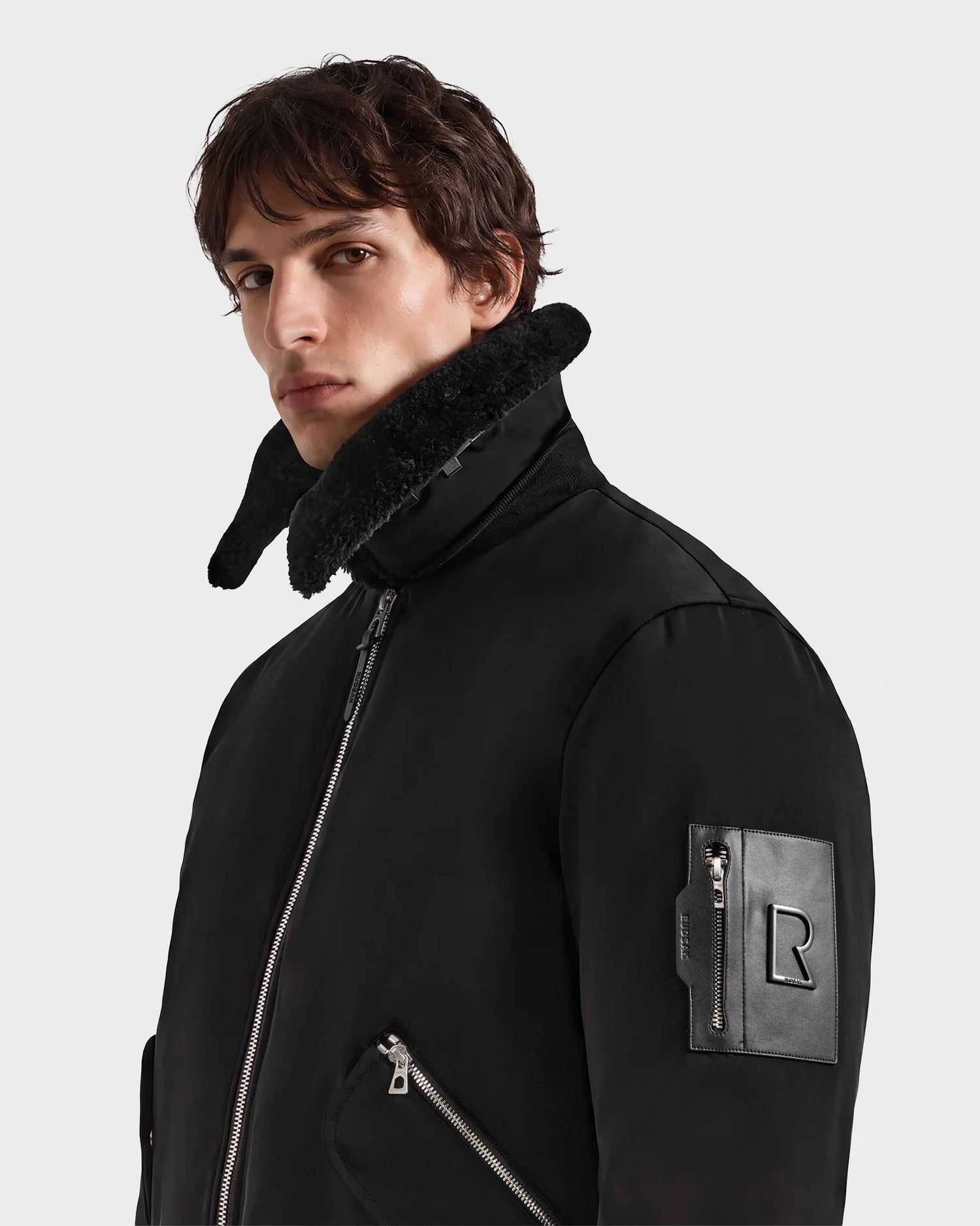 A person wearing the Rudsak DYLAN - 6125593 Black men's aviator bomber jacket, featuring a high faux fur collar and zippered pockets, stands against a plain background, looking slightly to the side—a perfect example of modern outerwear.