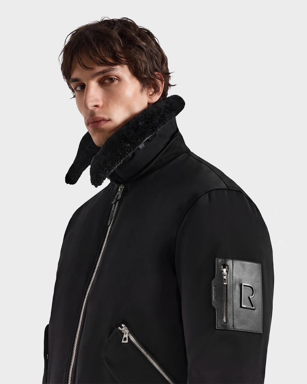 A person wearing the Rudsak DYLAN - 6125593 Black men's aviator bomber jacket, featuring a high faux fur collar and zippered pockets, stands against a plain background, looking slightly to the side—a perfect example of modern outerwear.