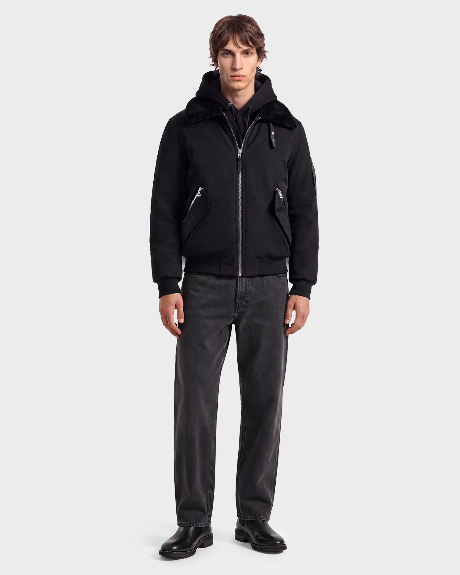 A man stands facing forward wearing the Rudsak DYLAN - 6125593 Black men’s aviator bomber jacket, featuring a faux fur collar, paired with a black hoodie, dark grey jeans, and black shoes against a plain light grey background. This modern outerwear look from Rudsak effortlessly combines style and comfort.
