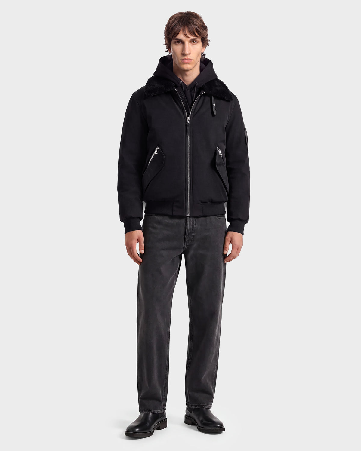 A man stands facing forward wearing the Rudsak DYLAN - 6125593 Black men’s aviator bomber jacket, featuring a faux fur collar, paired with a black hoodie, dark grey jeans, and black shoes against a plain light grey background. This modern outerwear look from Rudsak effortlessly combines style and comfort.