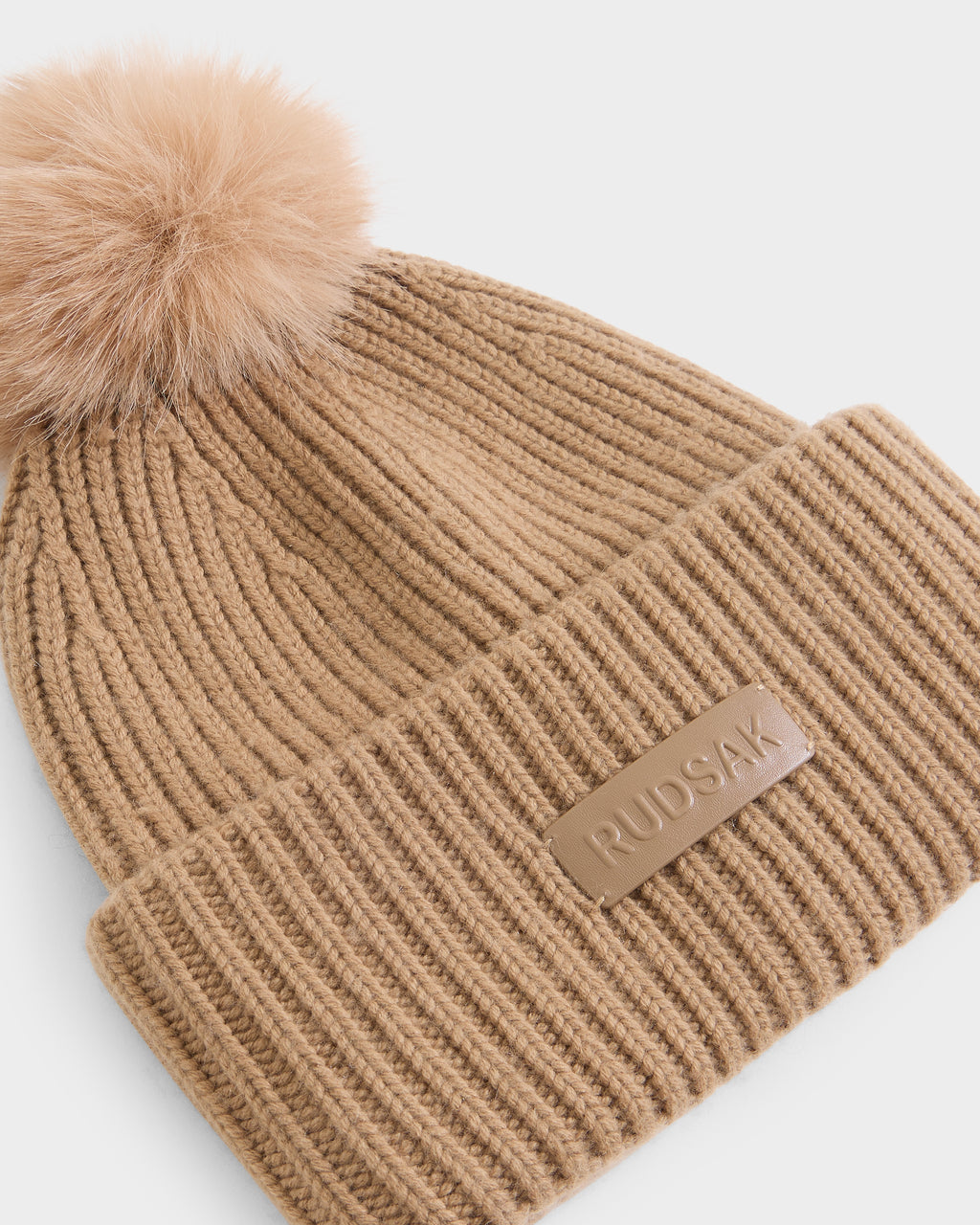 The DARA - 8425019 Camel by Rudsak is a stylish women’s winter hat in tan ribbed knit, featuring a large faux fur pom pom on top and a leather patch with the RUDSAK logo on the front cuff.