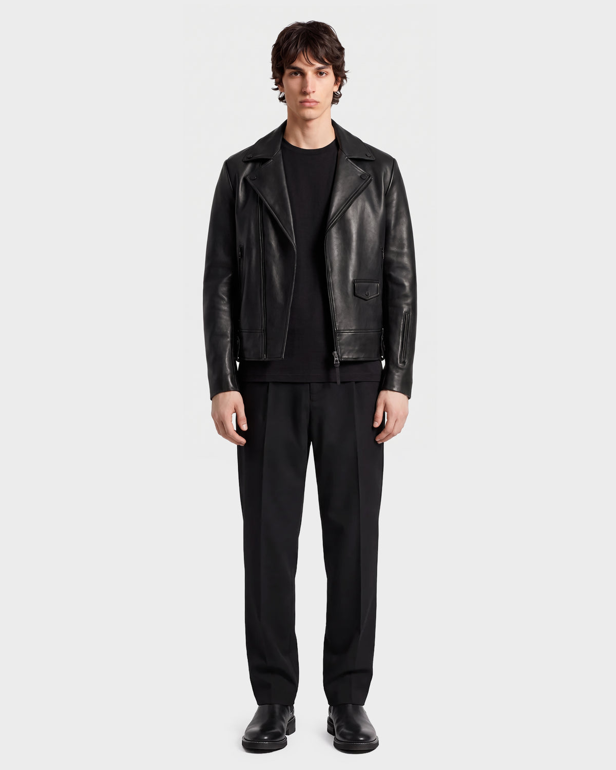 A person stands facing forward, wearing the RUDSAK DAREG - 6122004 BLACK lamb leather biker jacket—an undeniable wardrobe essential—paired with a black shirt, black trousers, and black shoes, against a plain light background.