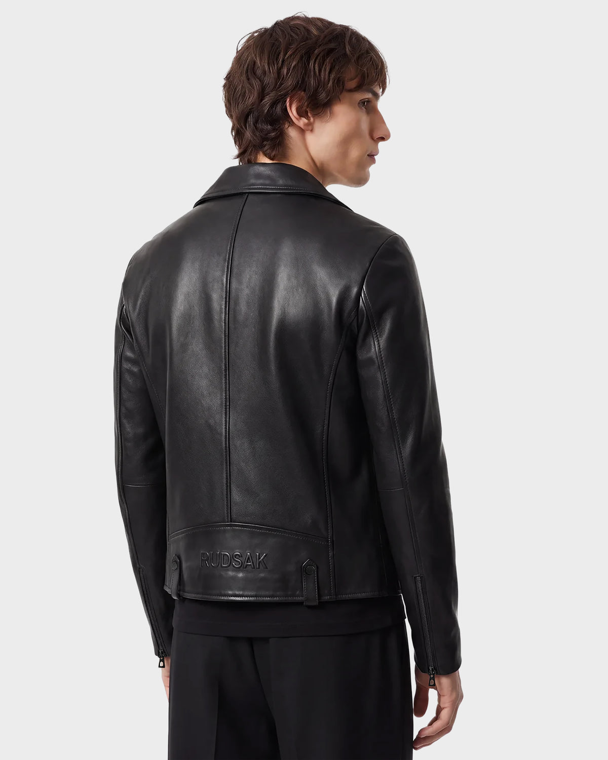 A person with short brown hair, facing away from the camera, is wearing the RUDSAK DAREG - 6122004 BLACK lamb leather biker jacket and black pants. The jacket—an essential piece—features "RUDSAK" embossed near the bottom on the back. The background is light gray.
